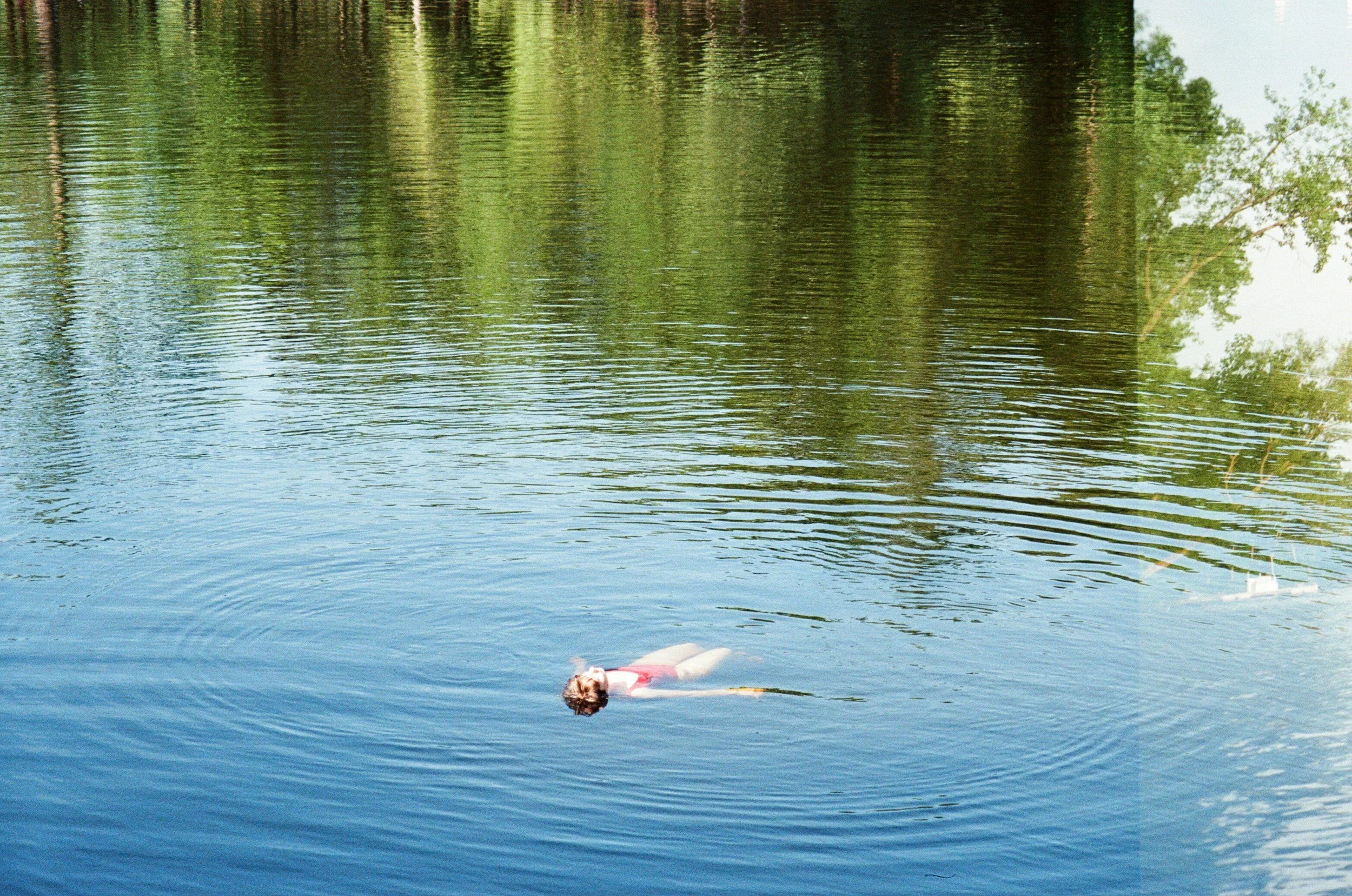 A person is floating on their back in a body of water, surrounded by trees and reflections on the water's surface.