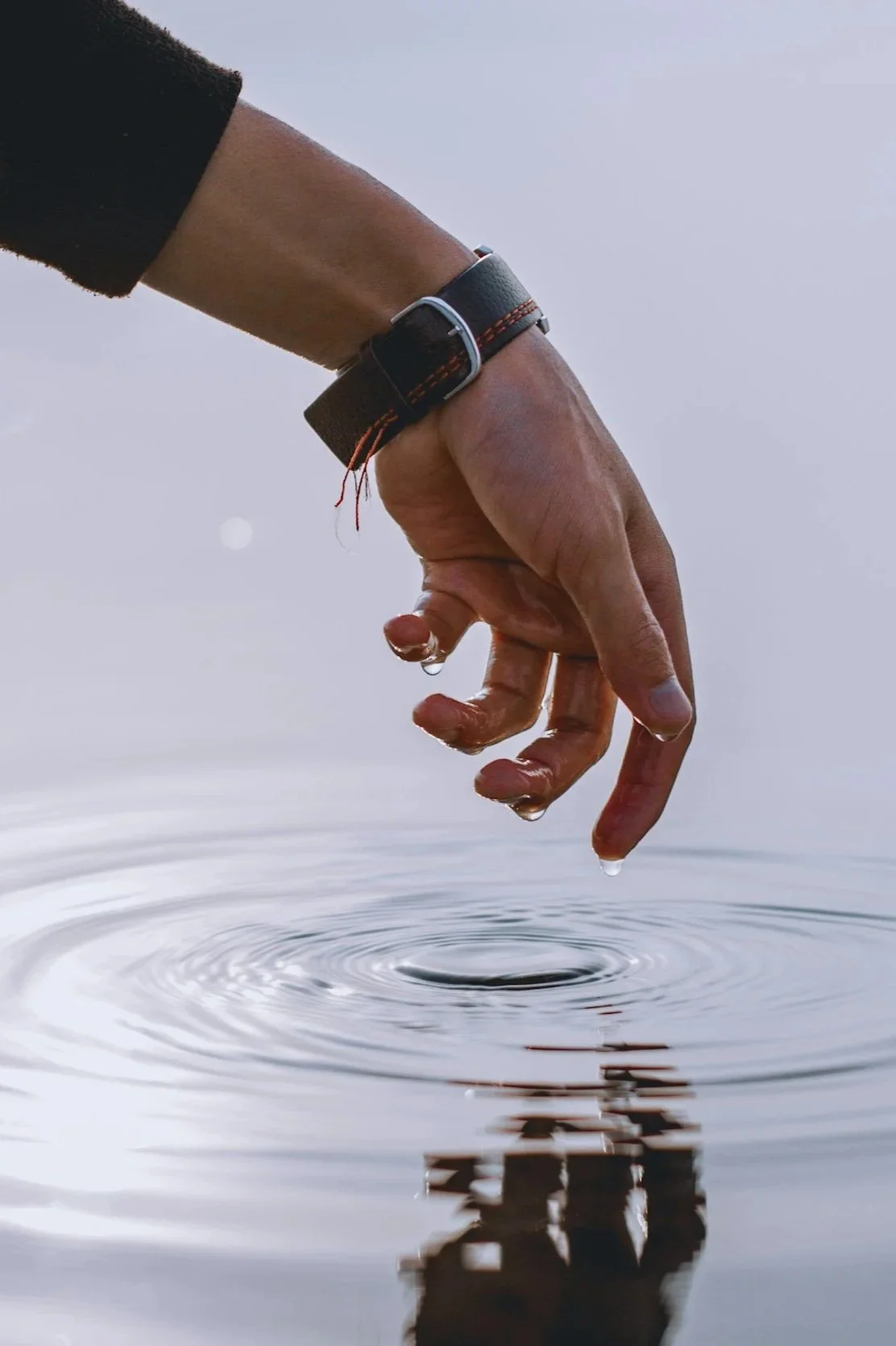 A person’s hand, wearing a black watch, touching water, creating ripples, with a few water droplets hanging from the fingers.