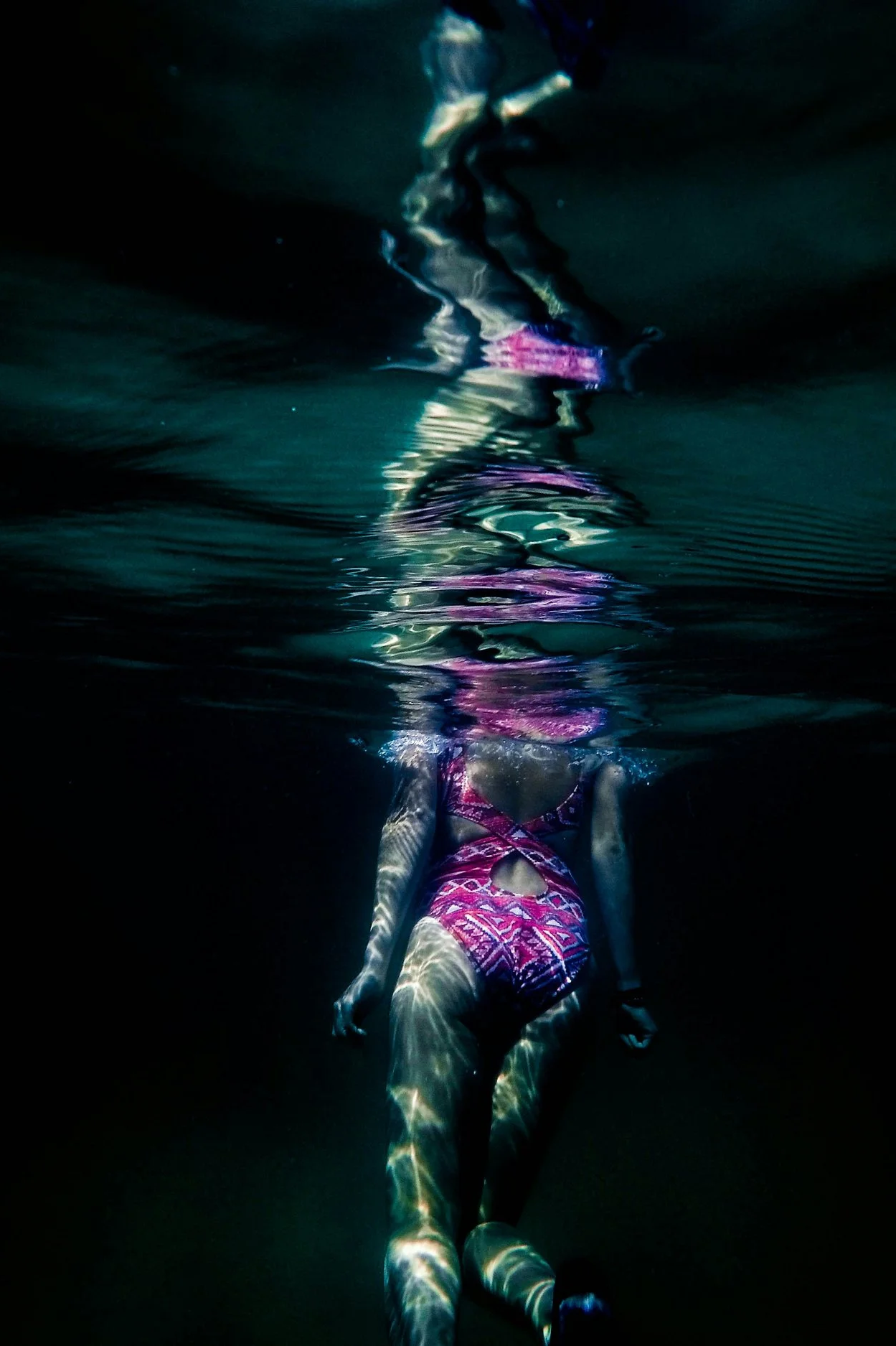 A person floating underwater wearing a colorful patterned swimsuit, with the surface water above and ripples creating light patterns on their body.