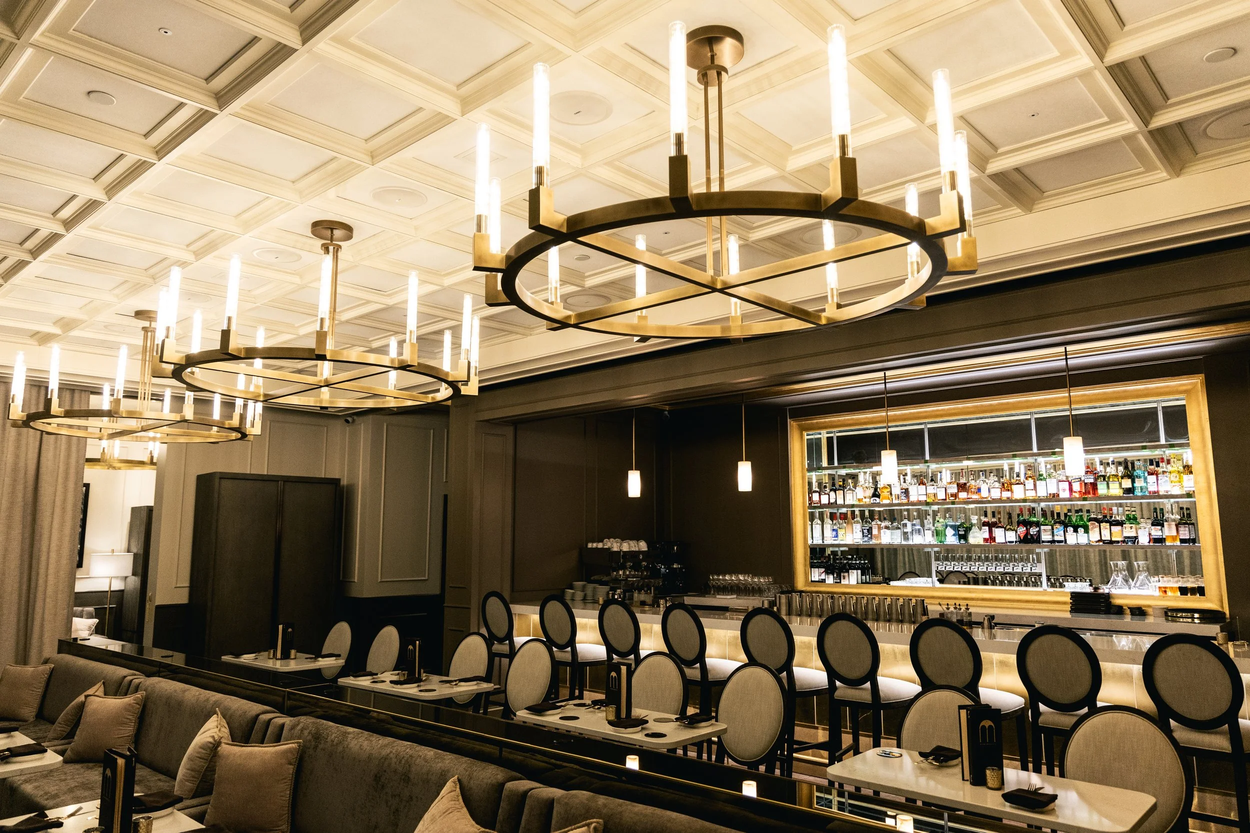 Elegant restaurant bar area with modern chandeliers and a well-stocked liquor shelf behind the bar.