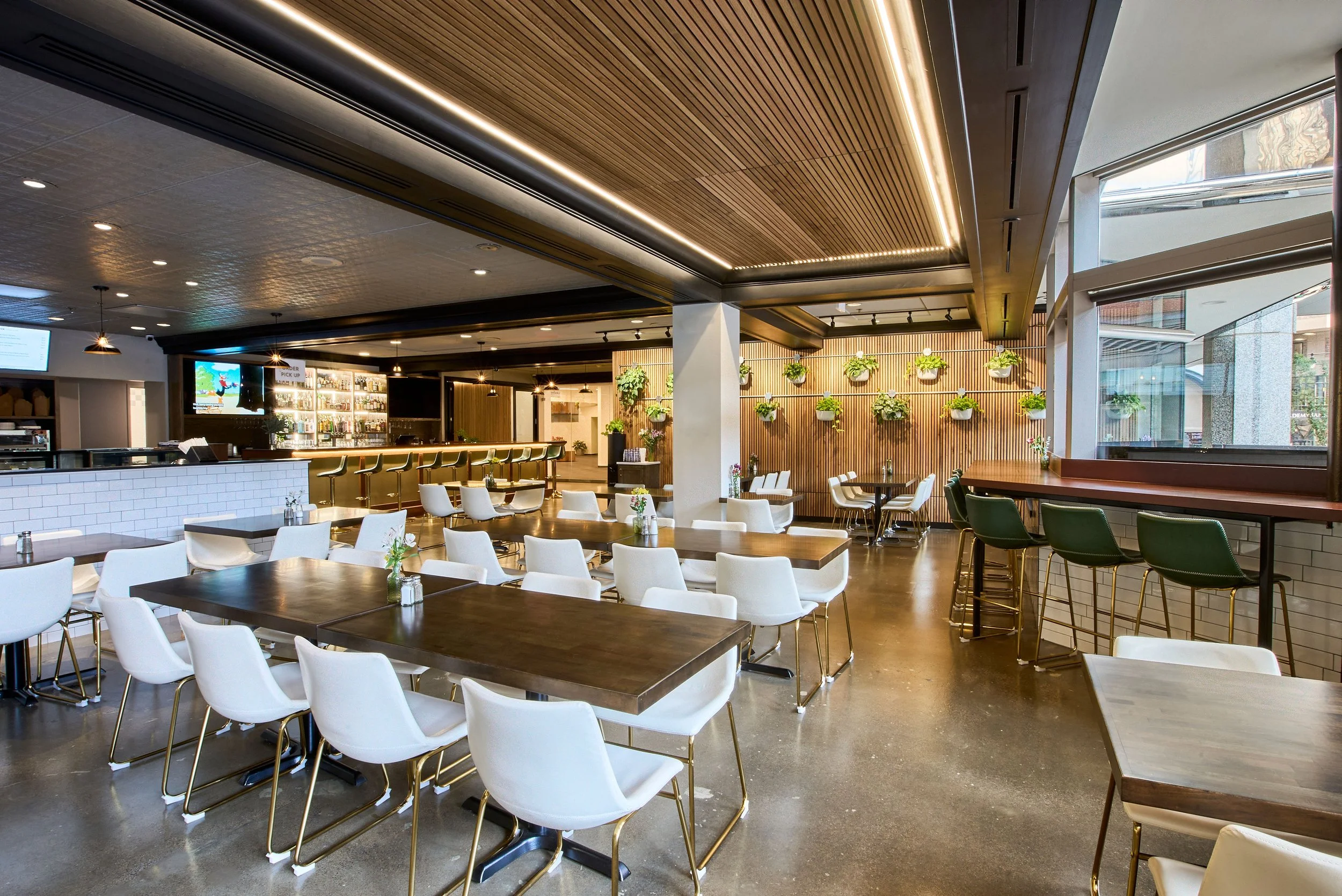 Modern restaurant interior with wooden ceiling, white and green chairs, dark wooden tables, and a bar with stools, decorated with plants and large windows.