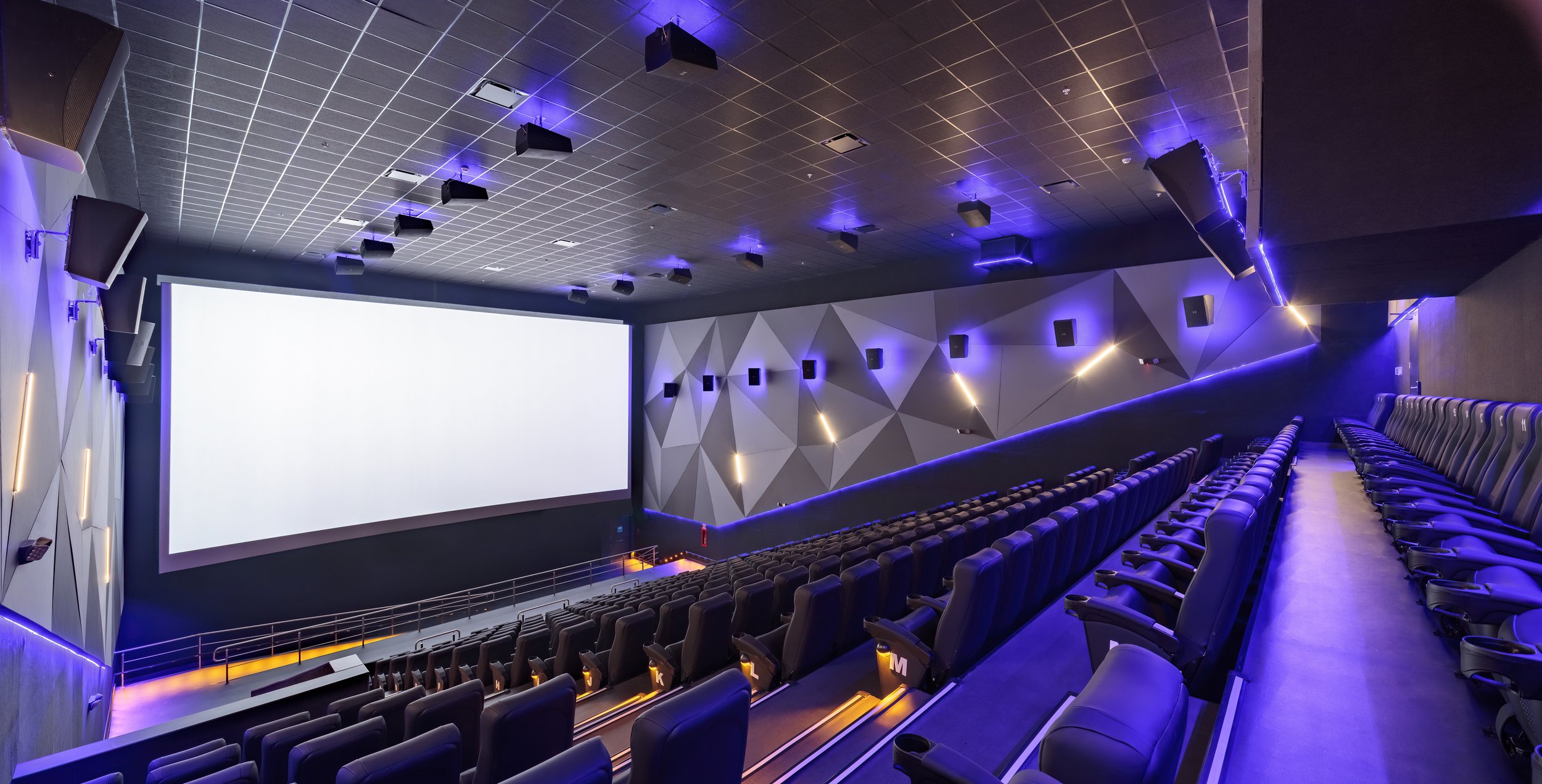 Empty modern movie theater with a large screen, multiple rows of black leather seats, purple and blue LED lighting, and sound speakers on the walls.