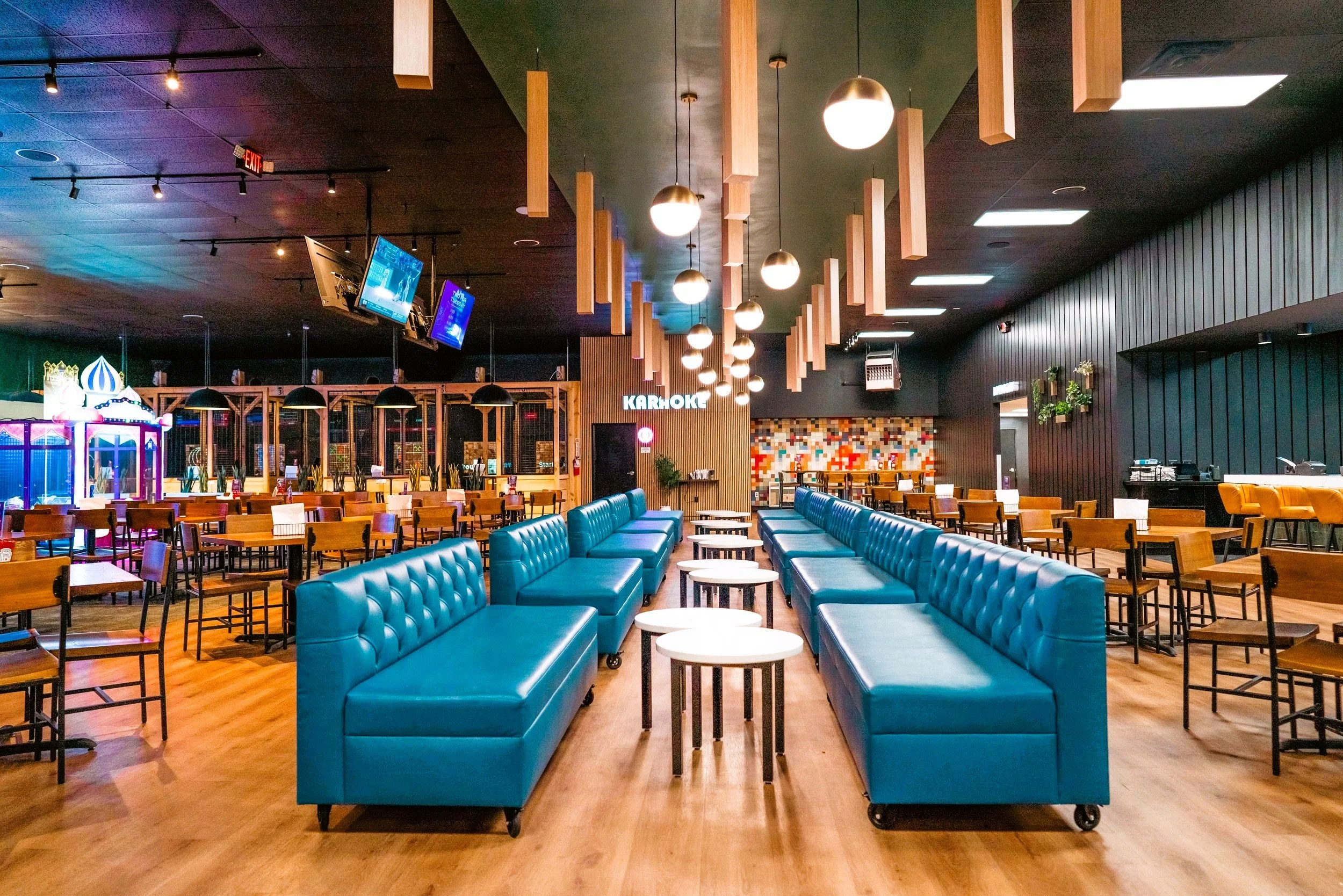 Interior of a karaoke bar with blue seating, wooden tables, a colorful wall in the background, and hanging lights.