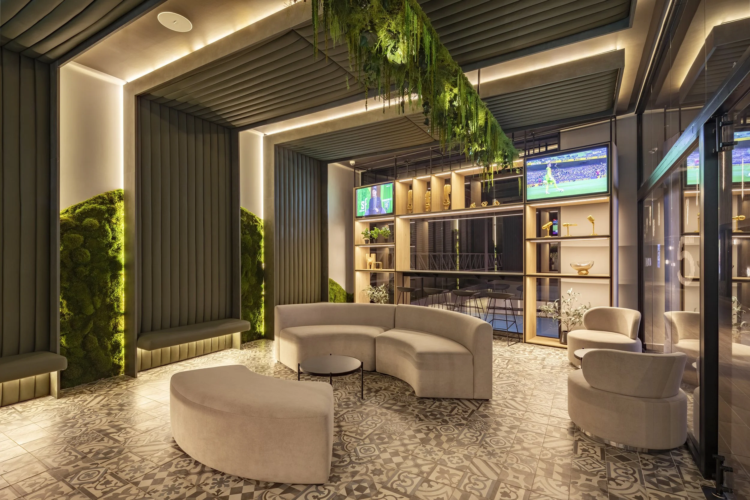 Modern lounge with beige curved sofa, armchairs, patterned tile floor, green moss wall, hanging greenery, wall-mounted screens displaying sports, and a wooden shelving unit with decorative items.