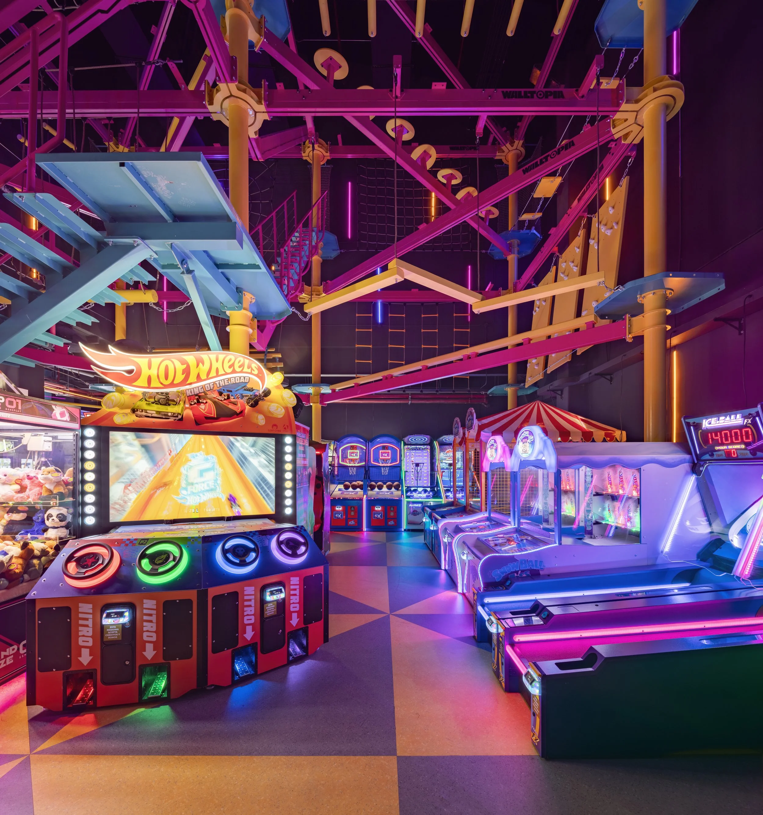 Colorful indoor arcade with neon lighting, featuring a Hot Wheels racing game, skee-ball machines, and an indoor climbing or play structure.