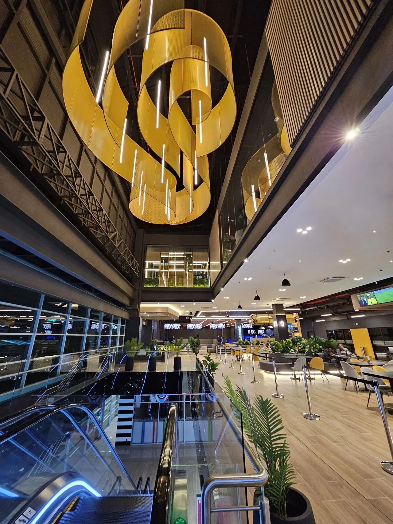 Interior view of a modern, upscale shopping mall or airport terminal with escalators, seating areas, plants, and a large gold spiral chandelier hanging from the ceiling.