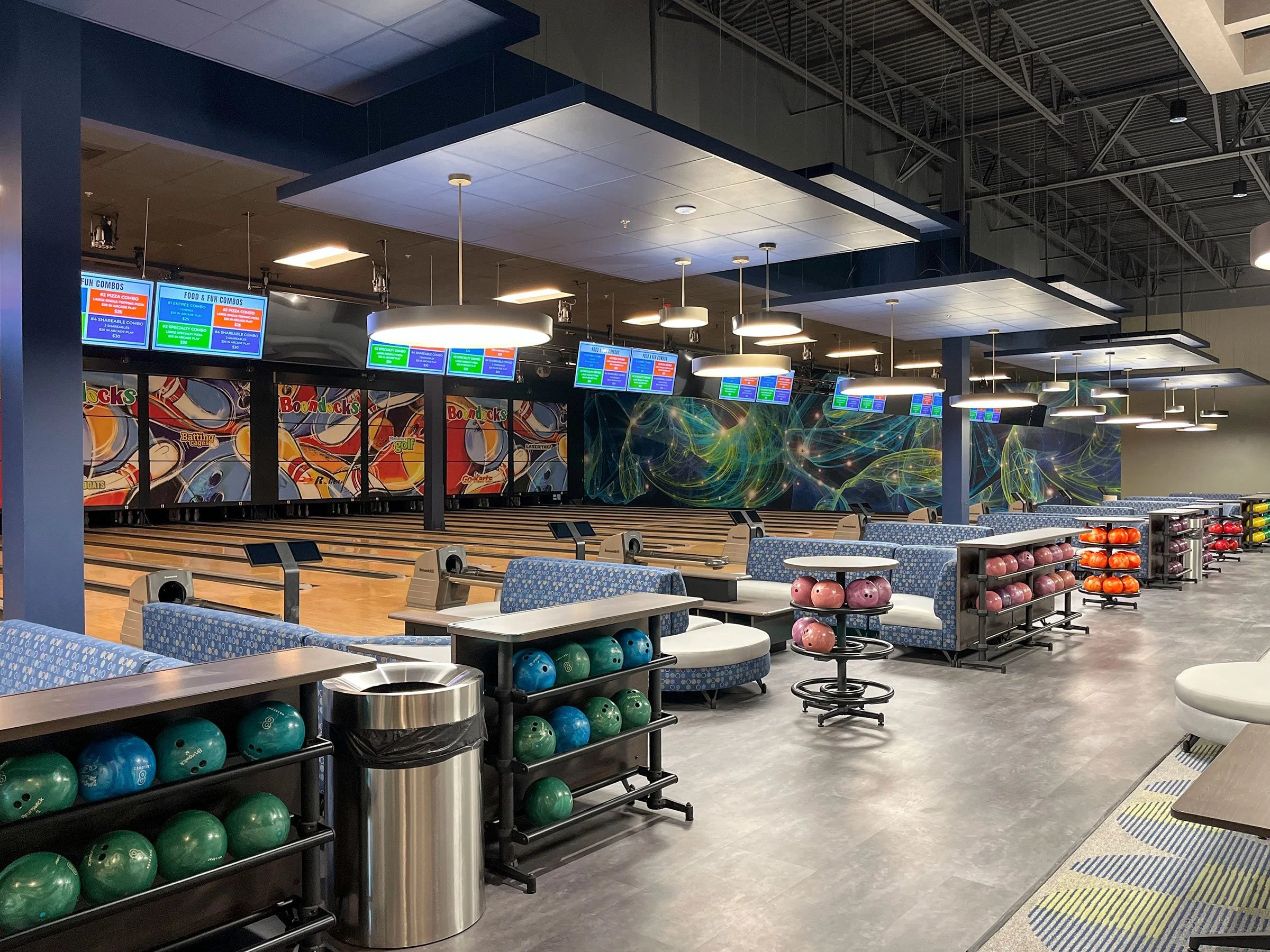 An empty bowling alley with multiple lanes, colorful bowling balls organized on racks, and digital screens displaying information above the lanes, with modern lighting and seating areas.