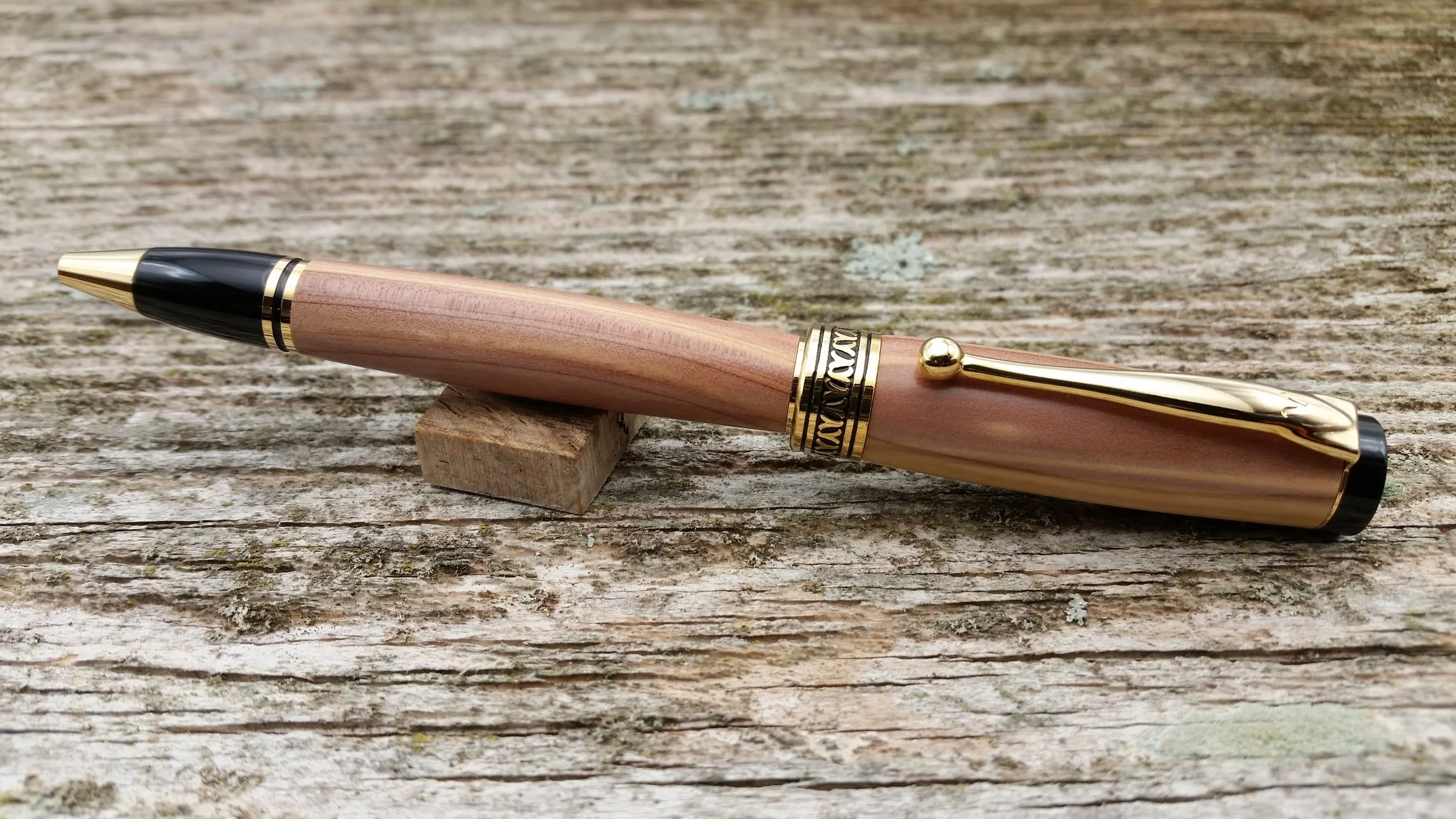 A ballpoint twist pen with a light wood body, gold accents, and a black tip, resting on a weathered wooden surface.