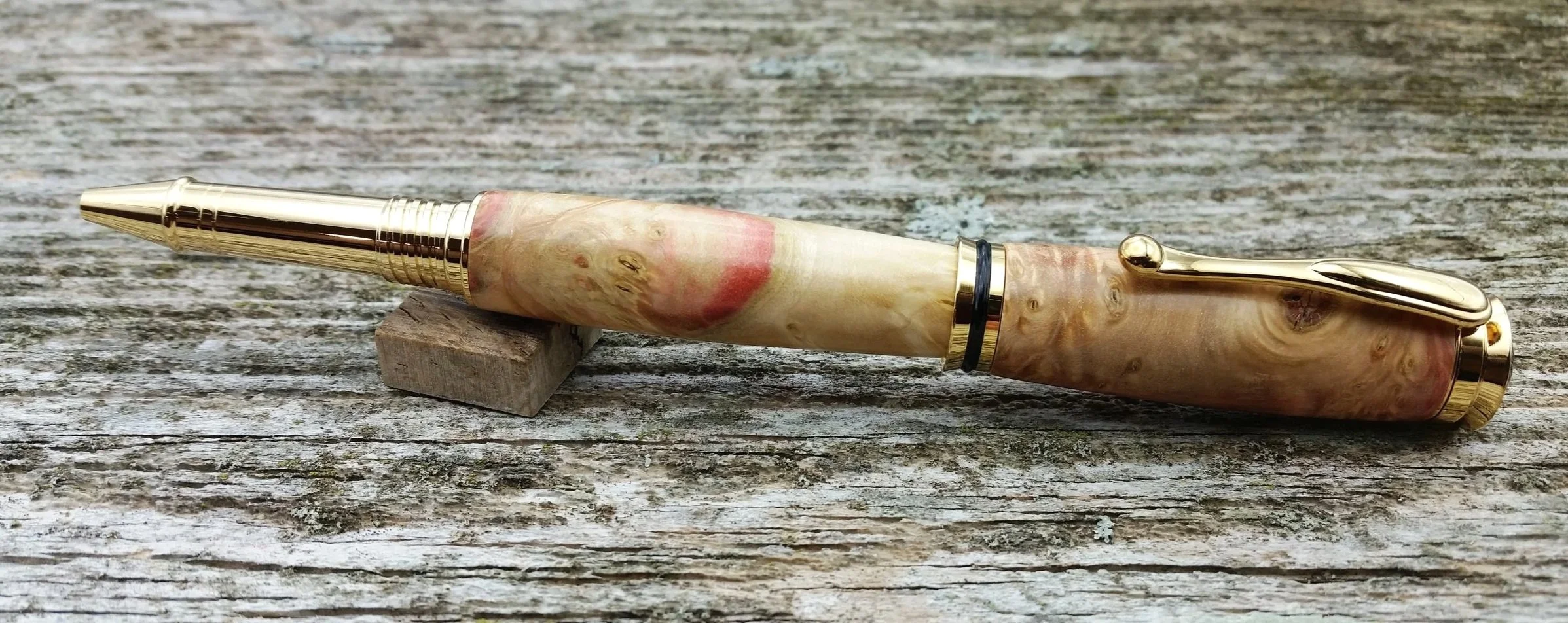 A rollerball pen with gold hardware and red flame box elder burl wood resting on a weathered wooden surface.