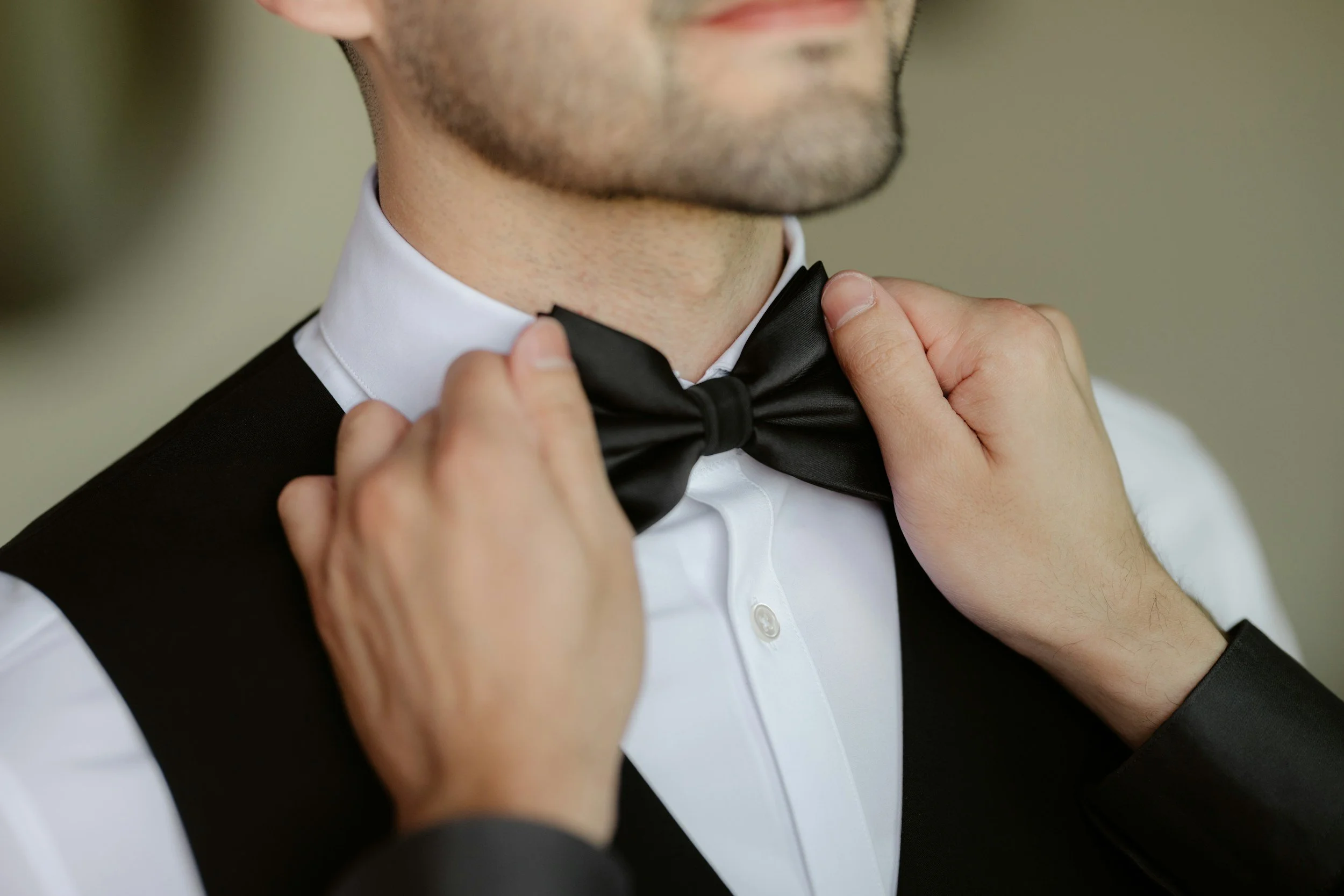 Person adjusting black bow tie while wearing a white shirt and black tuxedo vest.