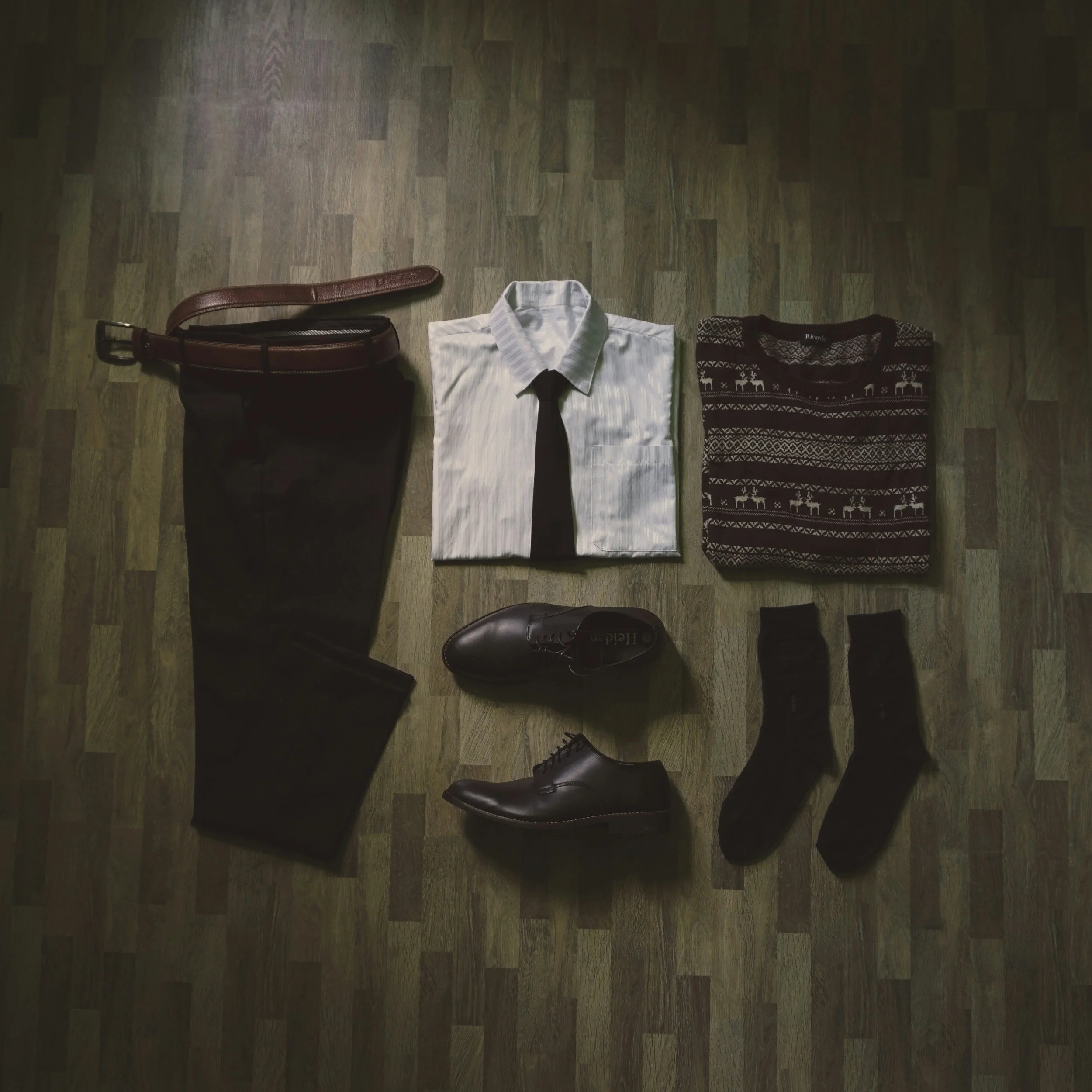 Men's dress outfit with black pants, brown belt, white shirt with a black tie, patterned sweater, black shoes, and black socks on wooden floor.