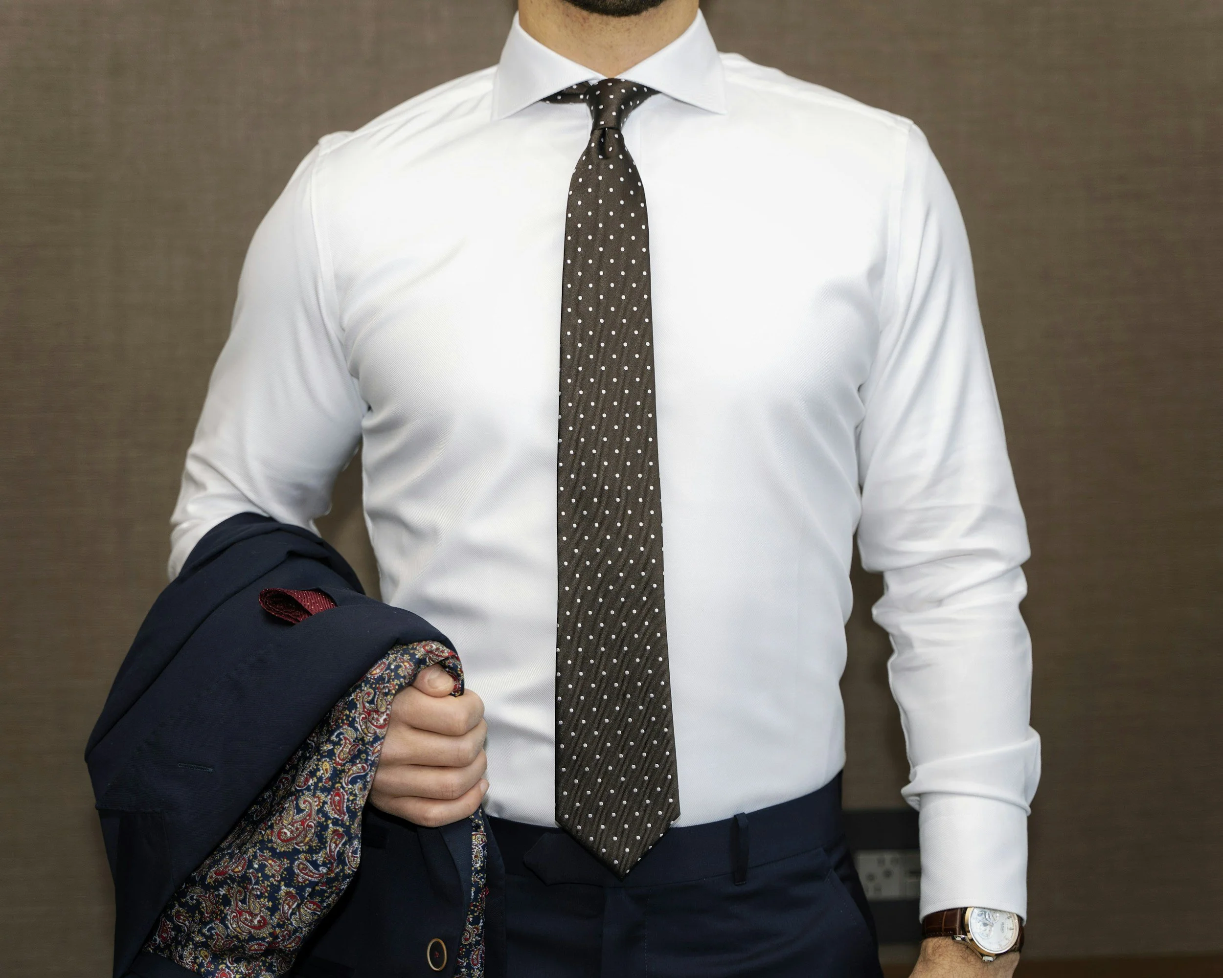 Man in a white dress shirt, dark polka dot tie, dark pants, holding a navy blazer with a patterned lining, and wearing a wristwatch