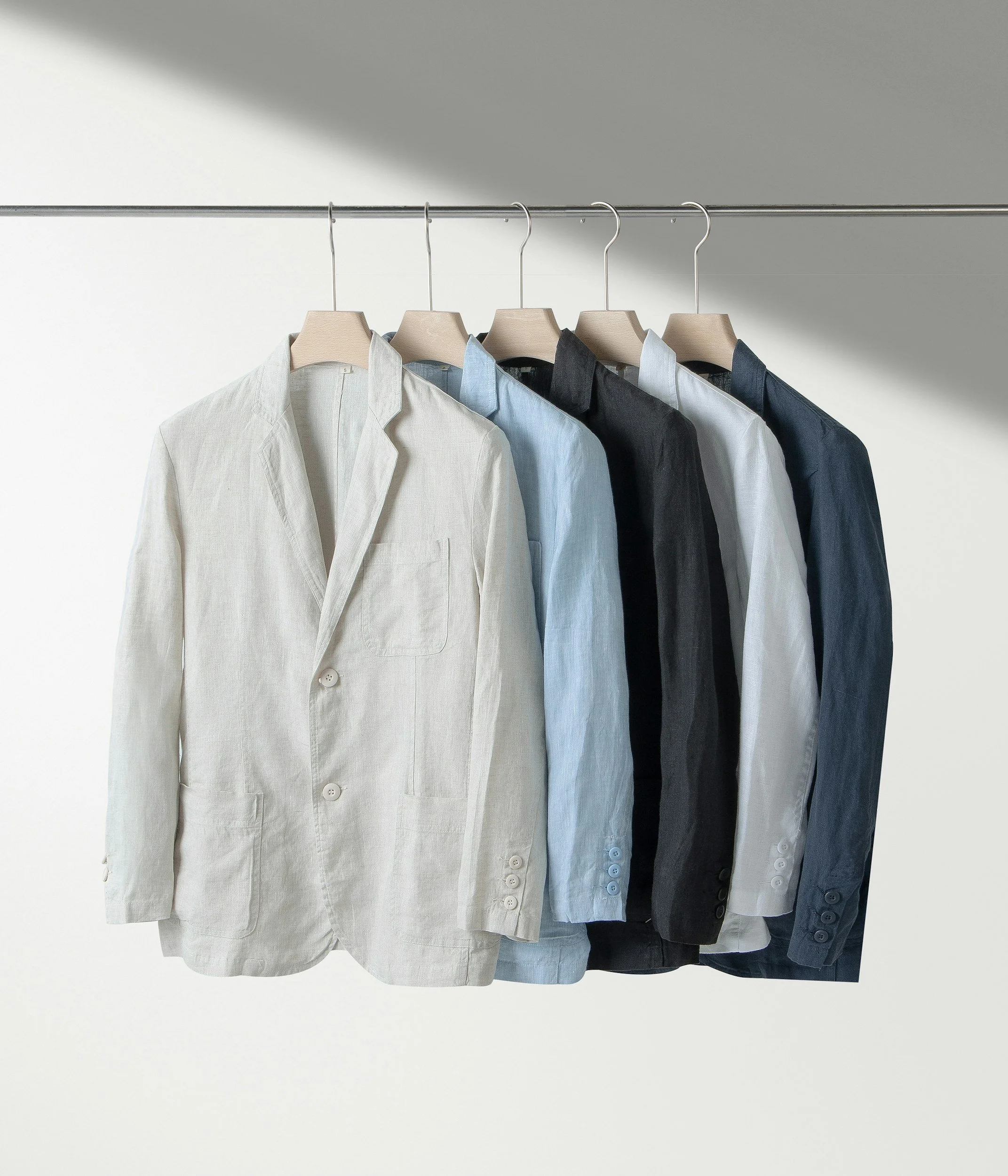 Five men's blazers hanging on a clothing rack, in colors ranging from white, light blue, black, white, to navy blue.