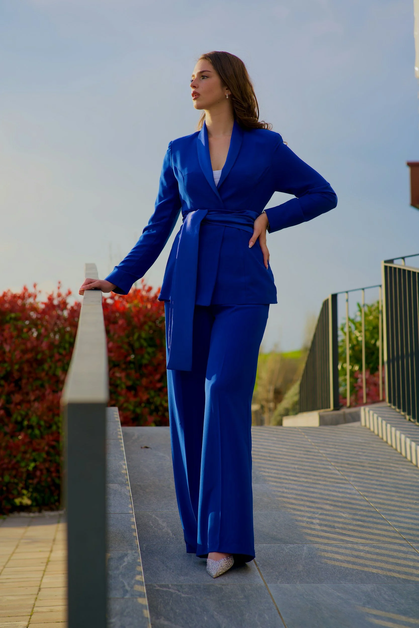 A woman in a bright blue suit with wide-leg pants and a belted blazer, standing outdoors on a ramp, looking into the distance with one hand resting on a rail and the other on her hip.