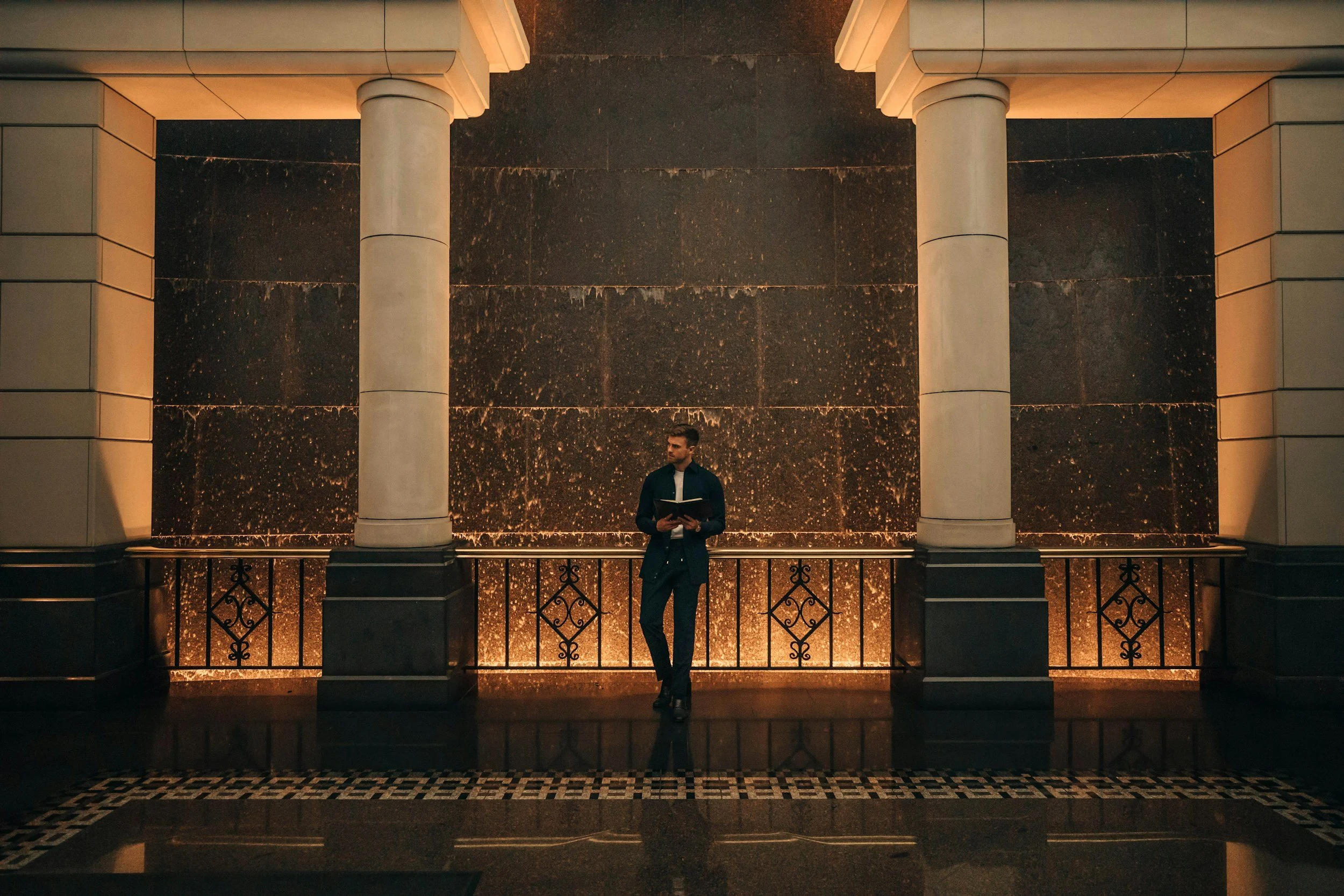 A man in a suit holding a folder leaning against a black ornate railing in front of a large, dark marble wall with warm lighting, inside a building with classical architectural elements. Montreal suot
