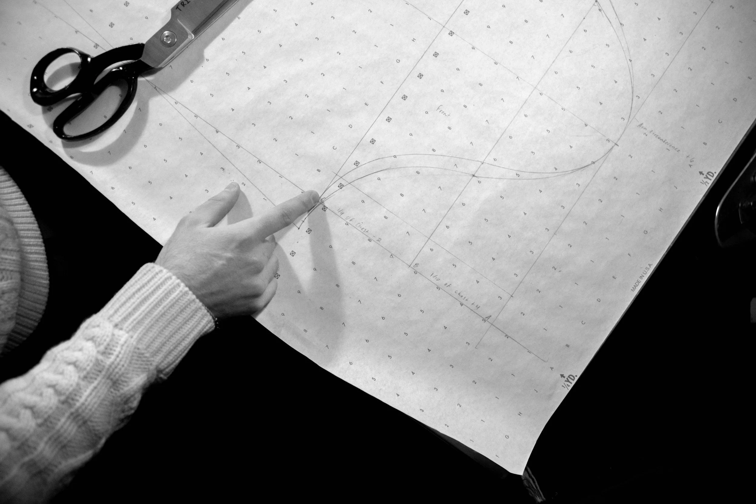 A person's hand pointing at a technical drawing on large paper, with black scissors and a cutting tool nearby, on a black surface.