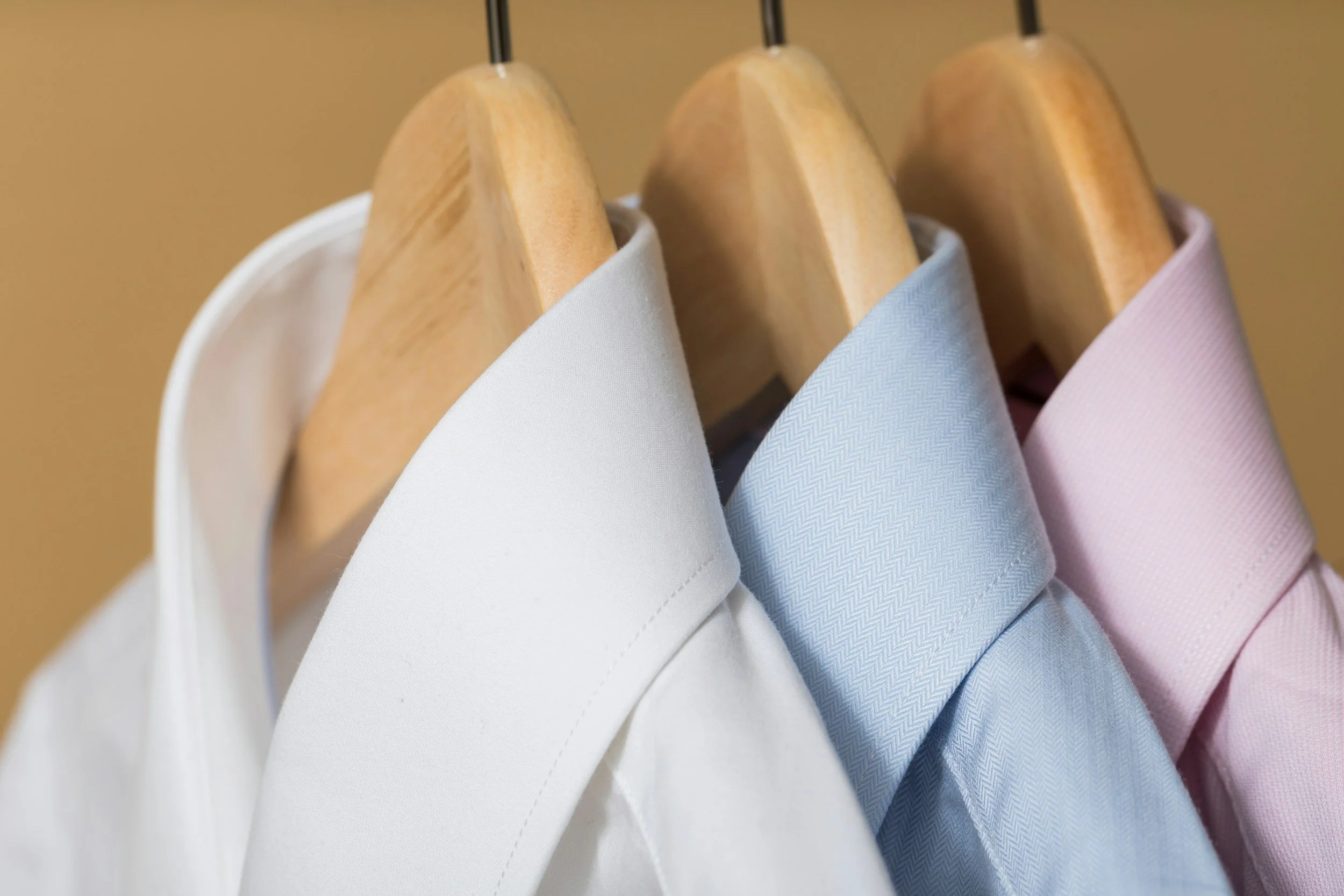 Three dress shirts in white, light blue, and pink hanging on wooden hangers.