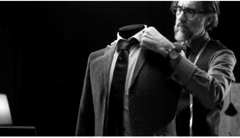 A man wearing glasses, a watch, and a shirt adjusts a suit on a mannequin in a dimly lit room.