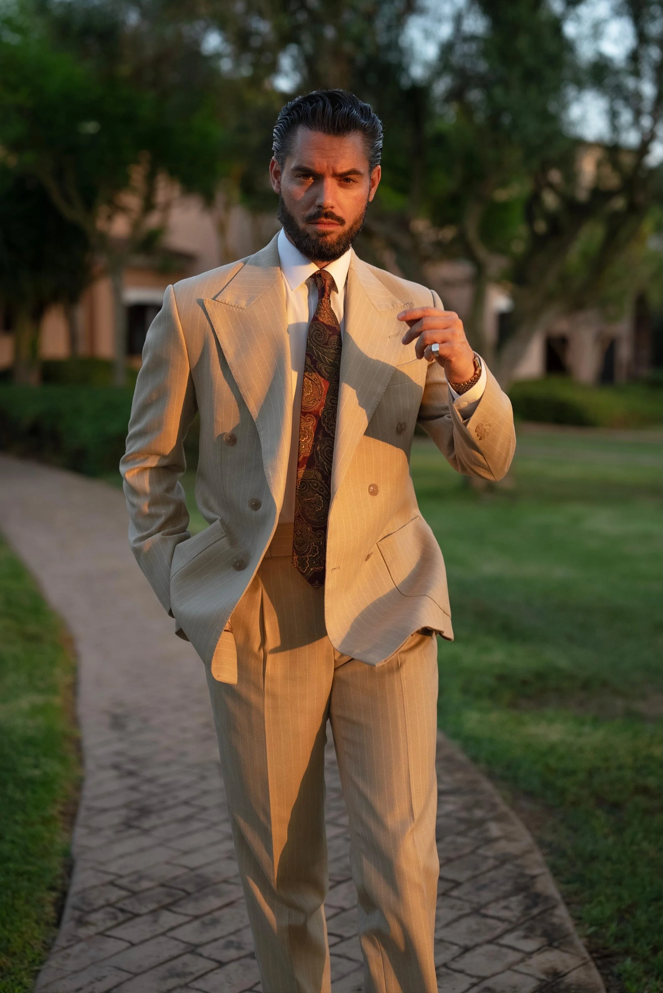 A man in a cream-colored pinstripe suit standing on a sidewalk outdoors during sunset, with trees and houses in the background.