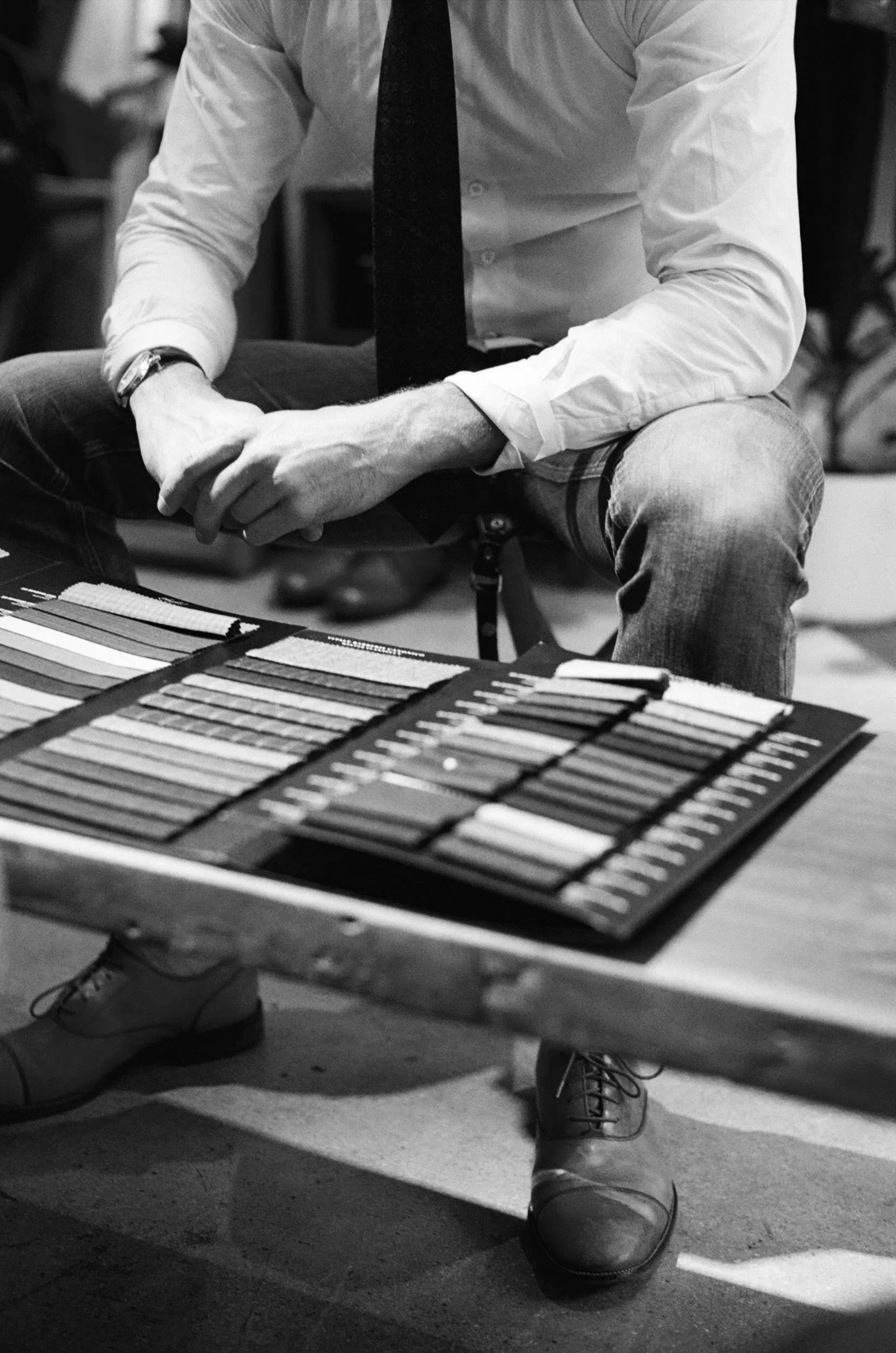 A man sitting at a table with fabric swatches, wearing a white shirt and a black tie, with only his torso and legs visible.