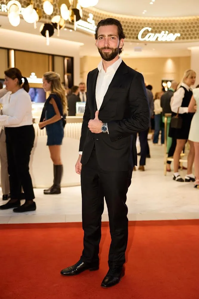 A man with a beard and dark hair wearing a black suit and white shirt, giving a thumbs-up, standing on a red carpet at a Cartier event inside a luxurious shopping mall or hotel, with other people and store displays in the background.