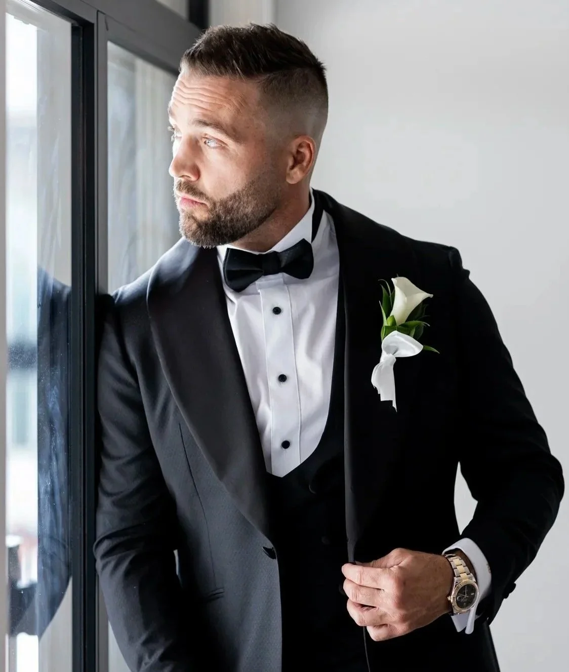 A man dressed in a black tuxedo with a bow tie and a white dress shirt, standing by a window with a boutonniere on his lapel, gazing outside.