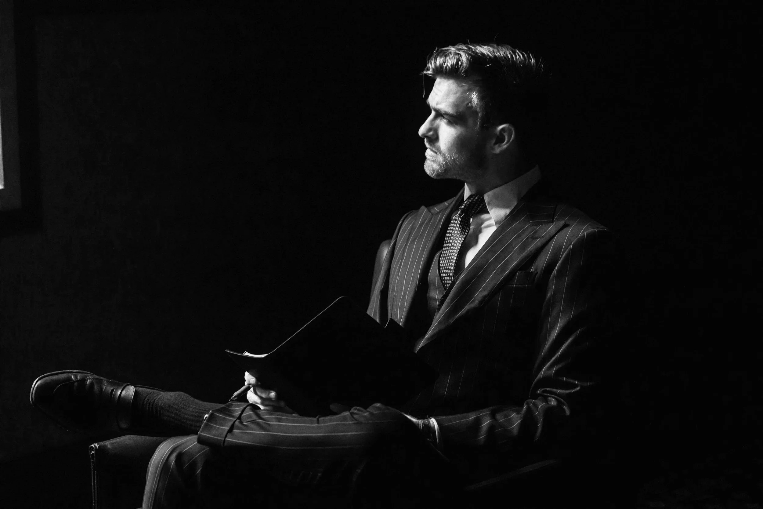 Black and white photo of a man in a pinstripe suit sitting with his legs crossed, holding a notebook, and looking to the left.