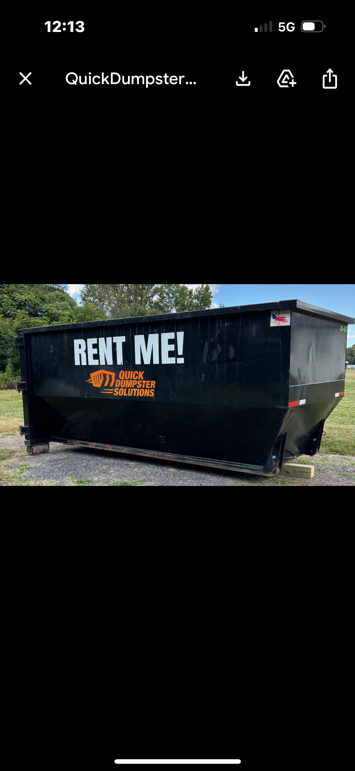 A black dumpster with the words "RENT ME!" in large white letters and the logo of Quick Dumpster Solutions in orange on the side, set outdoors on gravel with trees in the background.