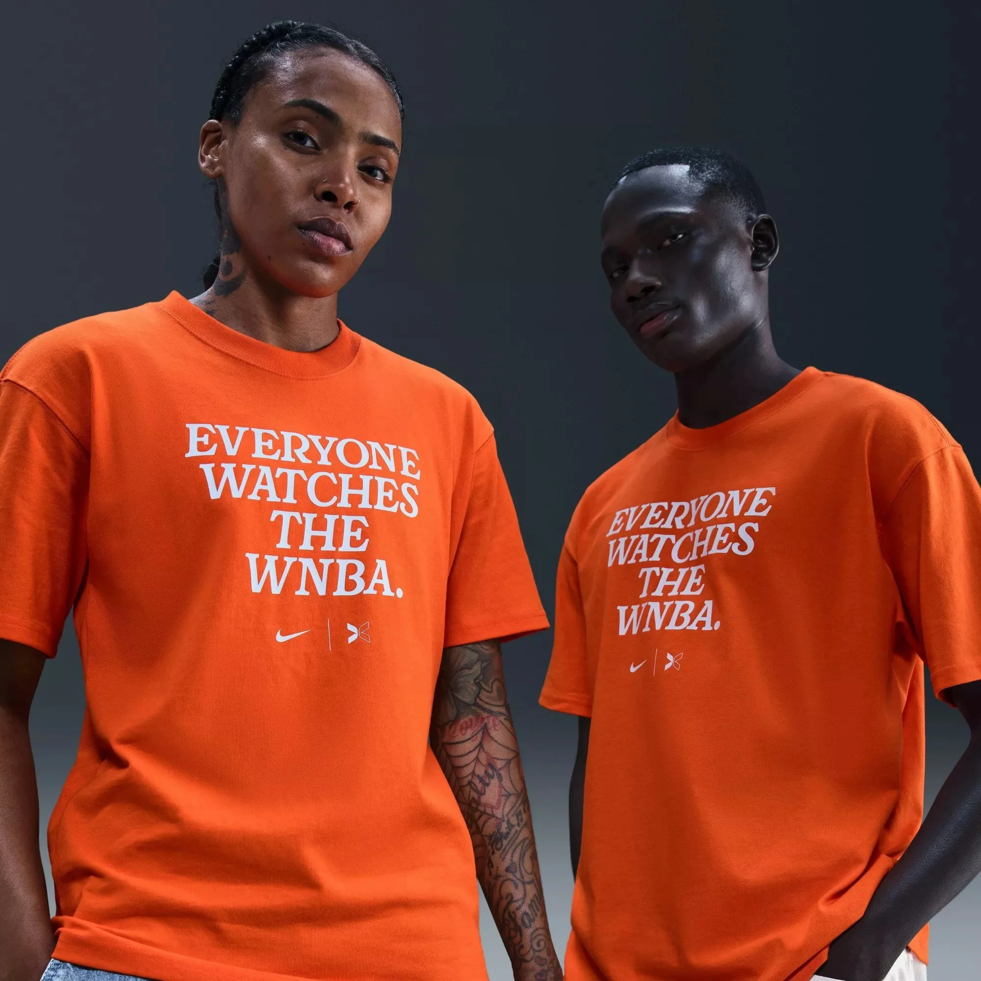 Two women wearing orange t-shirts with the phrase "Everyone Watches the WNBA" printed in white, standing close together against a dark background.