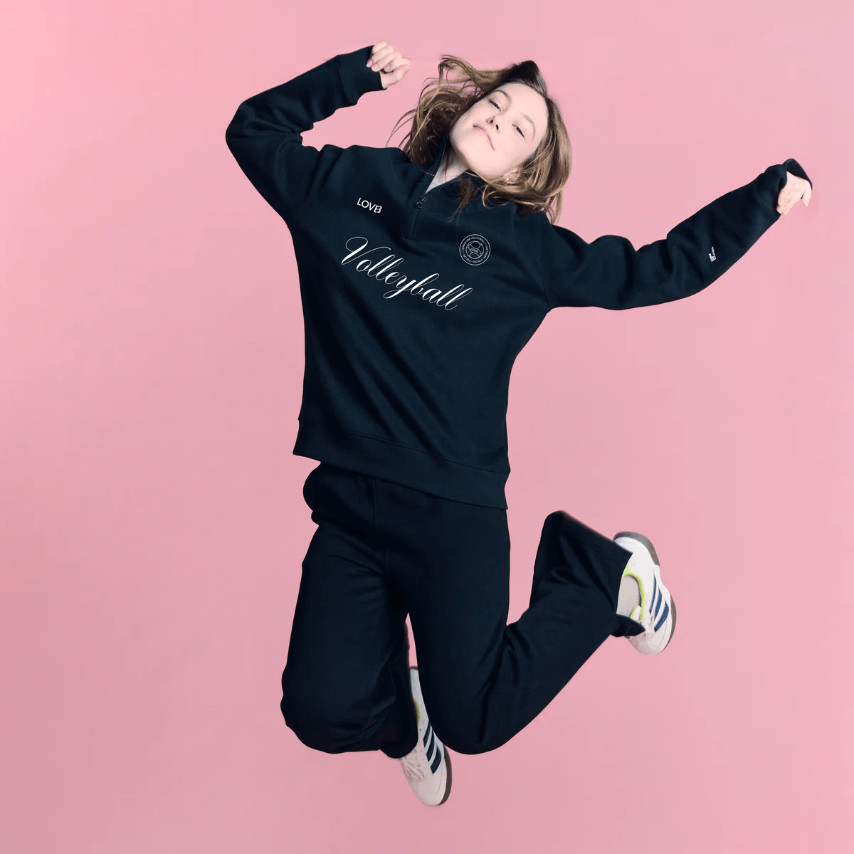A young woman jumping in the air against a pink background, wearing a black volleyball hoodie and black sweatpants, with white sneakers.