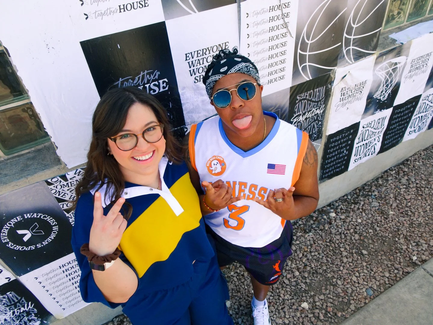 Two people posing for a photo outdoors in front of a wall decorated with posters. The woman on the left is smiling, wearing glasses and a blue and yellow jacket with a white collar, making a peace sign with her right hand. The man on the right is sticking out his tongue, wearing sunglasses, a bandana, a white sports jersey with orange and blue accents, and making a hand gesture. The ground is covered with small rocks.