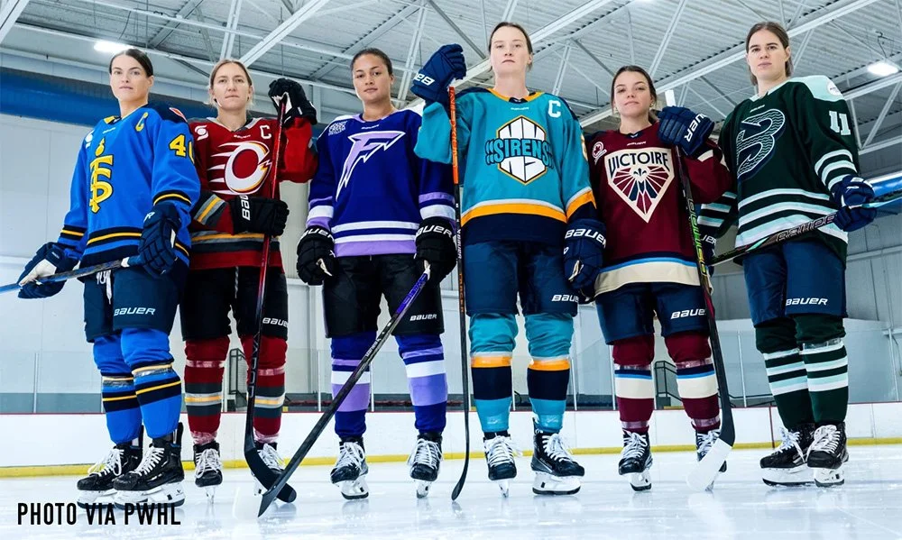 Six female ice hockey players standing on the ice rink in their team uniforms, holding hockey sticks, inside an indoor ice arena.