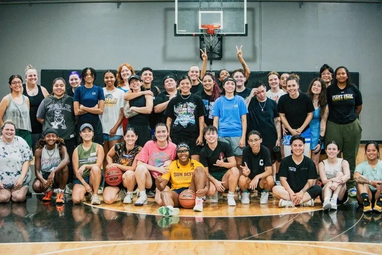 Group of women and girls in a gymnasium, some holding basketballs, posing for a photo in front of a basketball hoop, smiling and making peace signs.