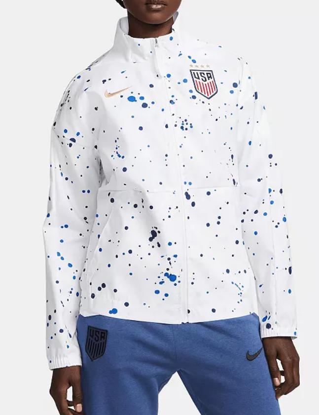 A sports jacket with the USA crest on the upper left chest, a Nike logo on the right chest, and a splatter paint design in blue, black, and navy on a white background, worn by a woman.