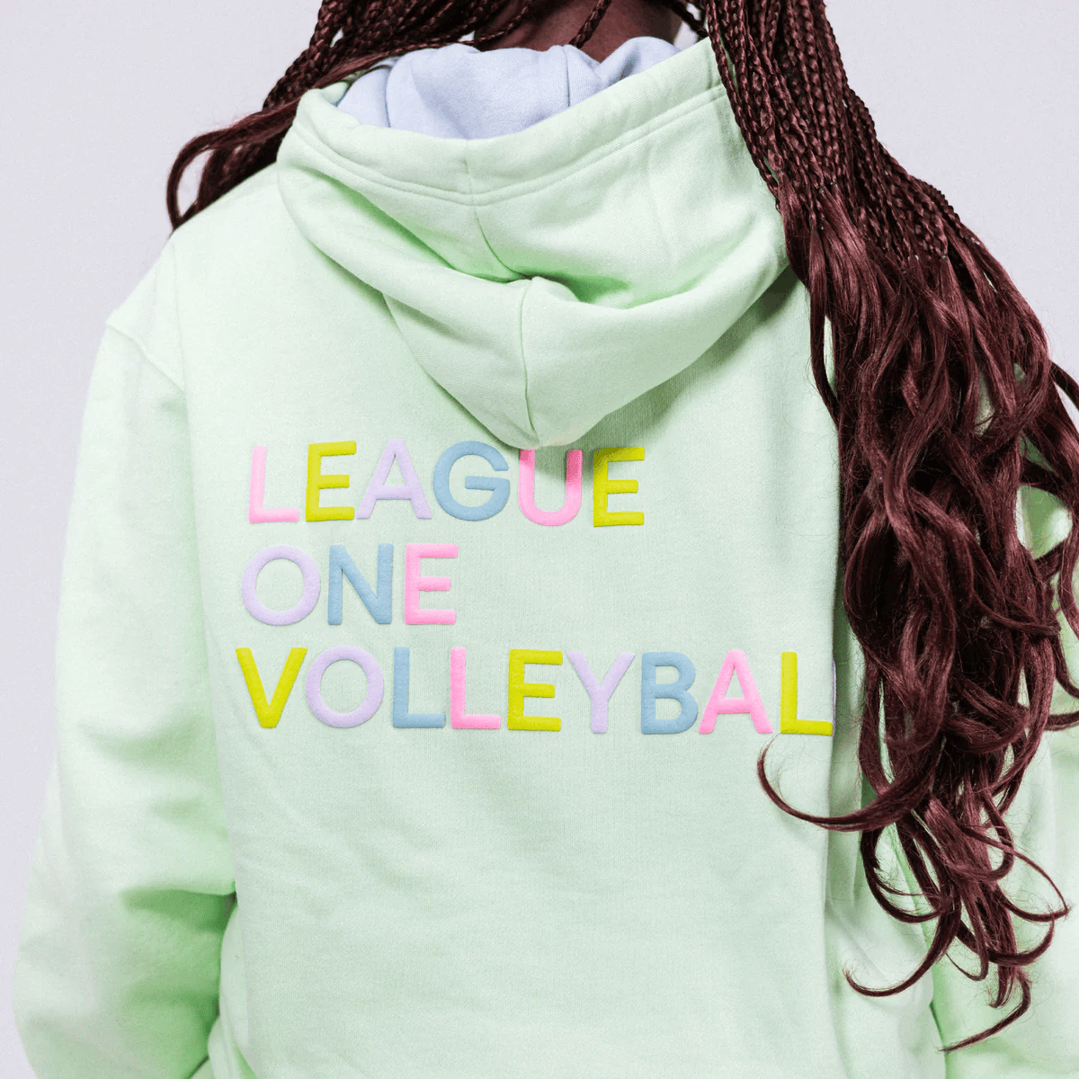 Back of a person wearing a light green hoodie with colorful text that reads "LEAGUE ONE VOLLEYBALL" and long, dark red braided hair.