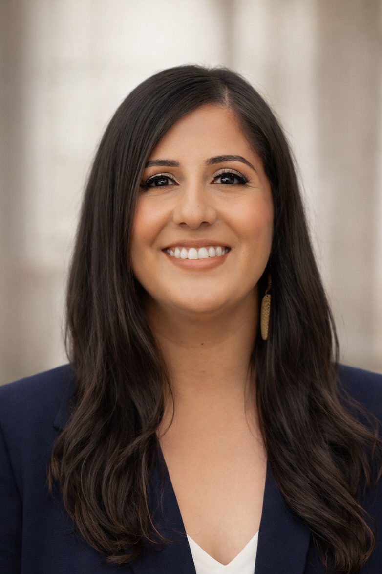 A woman with long dark hair, wearing makeup and a navy blazer, smiling at the camera with a blurred neutral background.