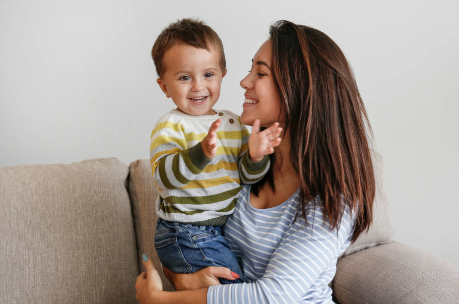 A woman with brown hair holding a smiling young boy with curly hair on her lap, sitting on a beige couch in a living room with a plain white wall background.