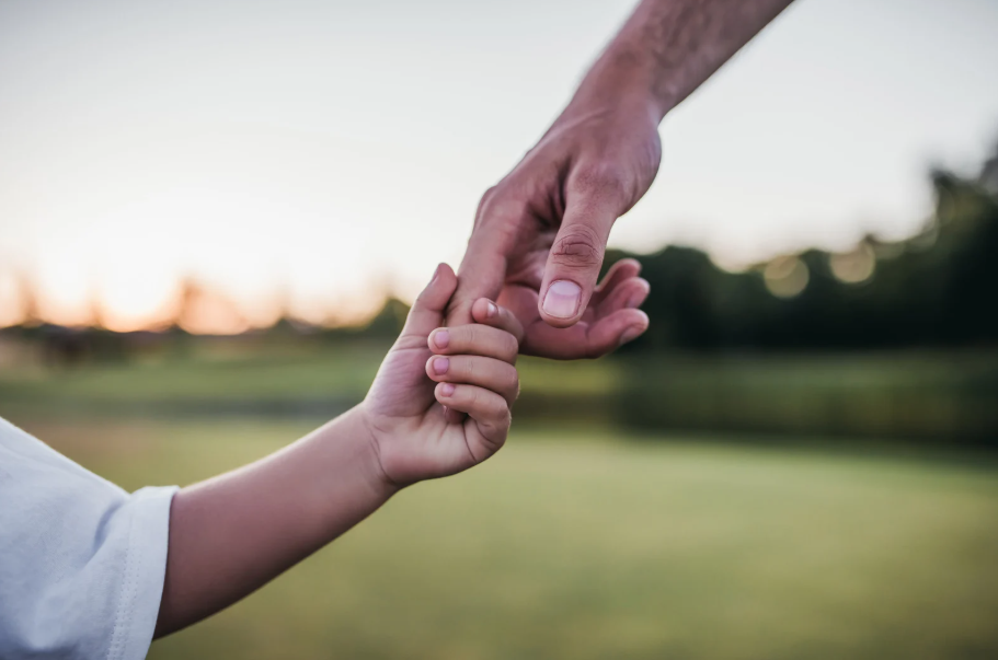 An adult's hand gently holding a child's hand outdoors during sunset or sunrise.