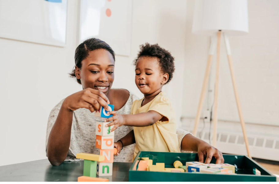 A woman and a young girl playing with colorful alphabet blocks at a table in a bright room.