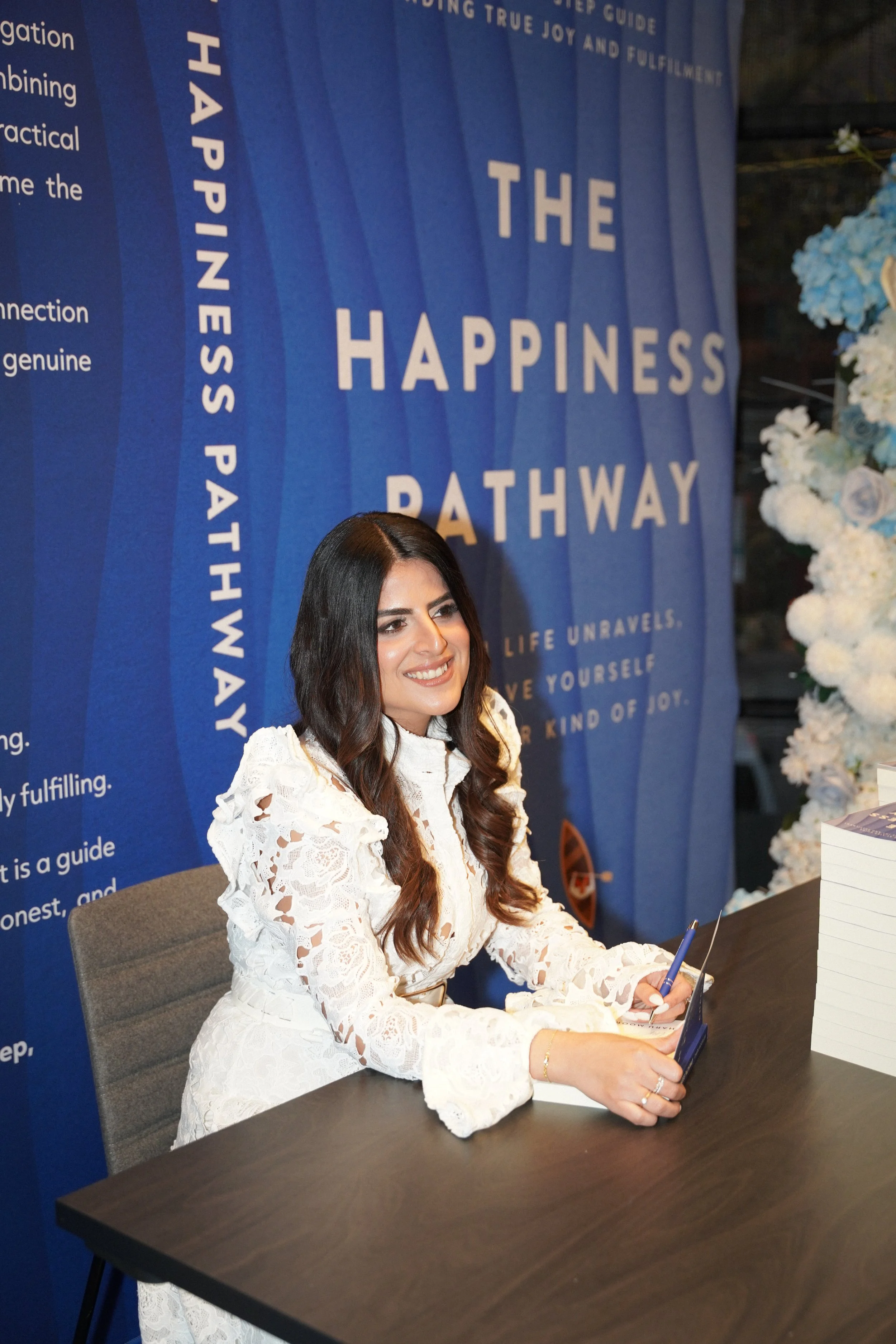 A smiling woman with long dark hair sitting at a black table, holding a pen, in front of a blue promotional backdrop for a happiness pathway event. The woman is dressed in a white lace dress.