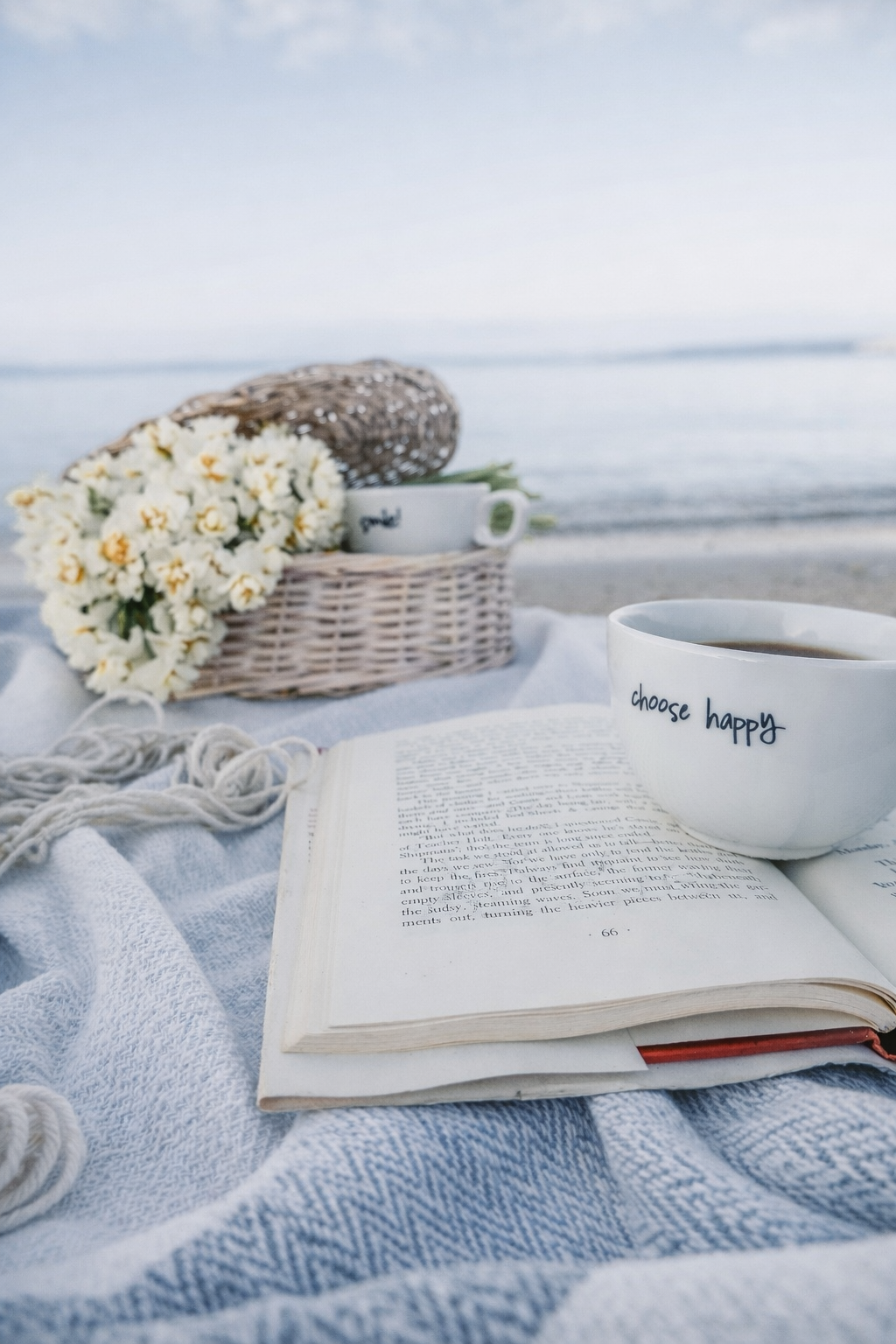 A cozy beach scene with a book, a mug that says 'choose happy', a basket of white flowers, and a cup of coffee on a blanket by the ocean.