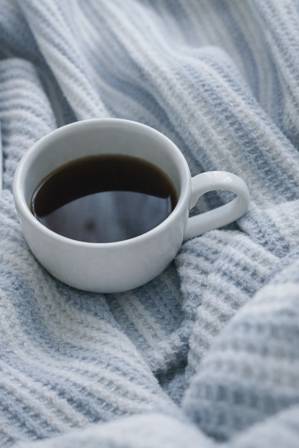 A white mug filled with black coffee resting on a soft, gray, striped blanket.