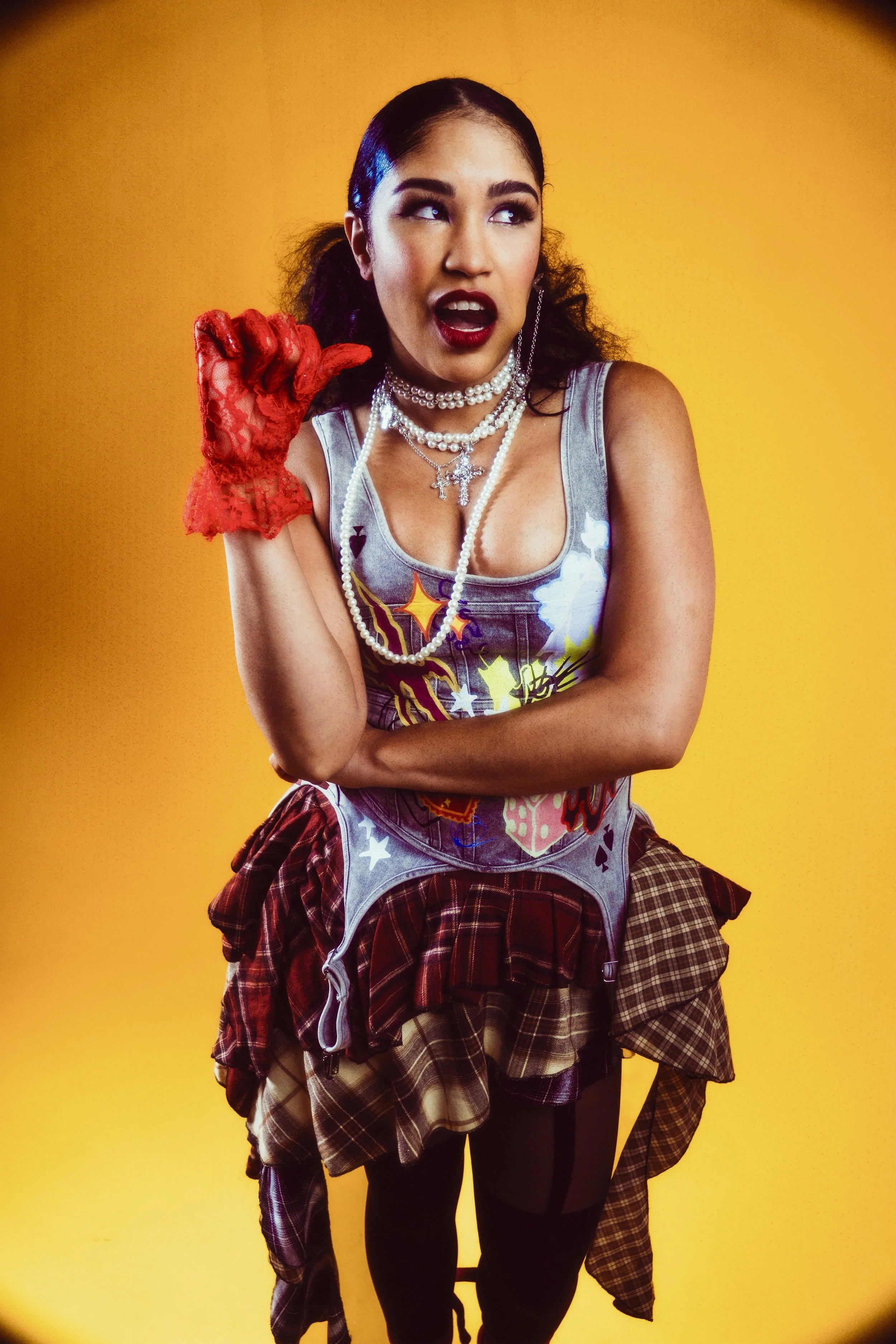 Young woman with dark hair in pigtails, wearing makeup, a pearl necklace, a sleeveless shirt with colorful patterns, a plaid skirt, tights, and a red lace glove, against a yellow background, making a playful gesture with her hand near her face.