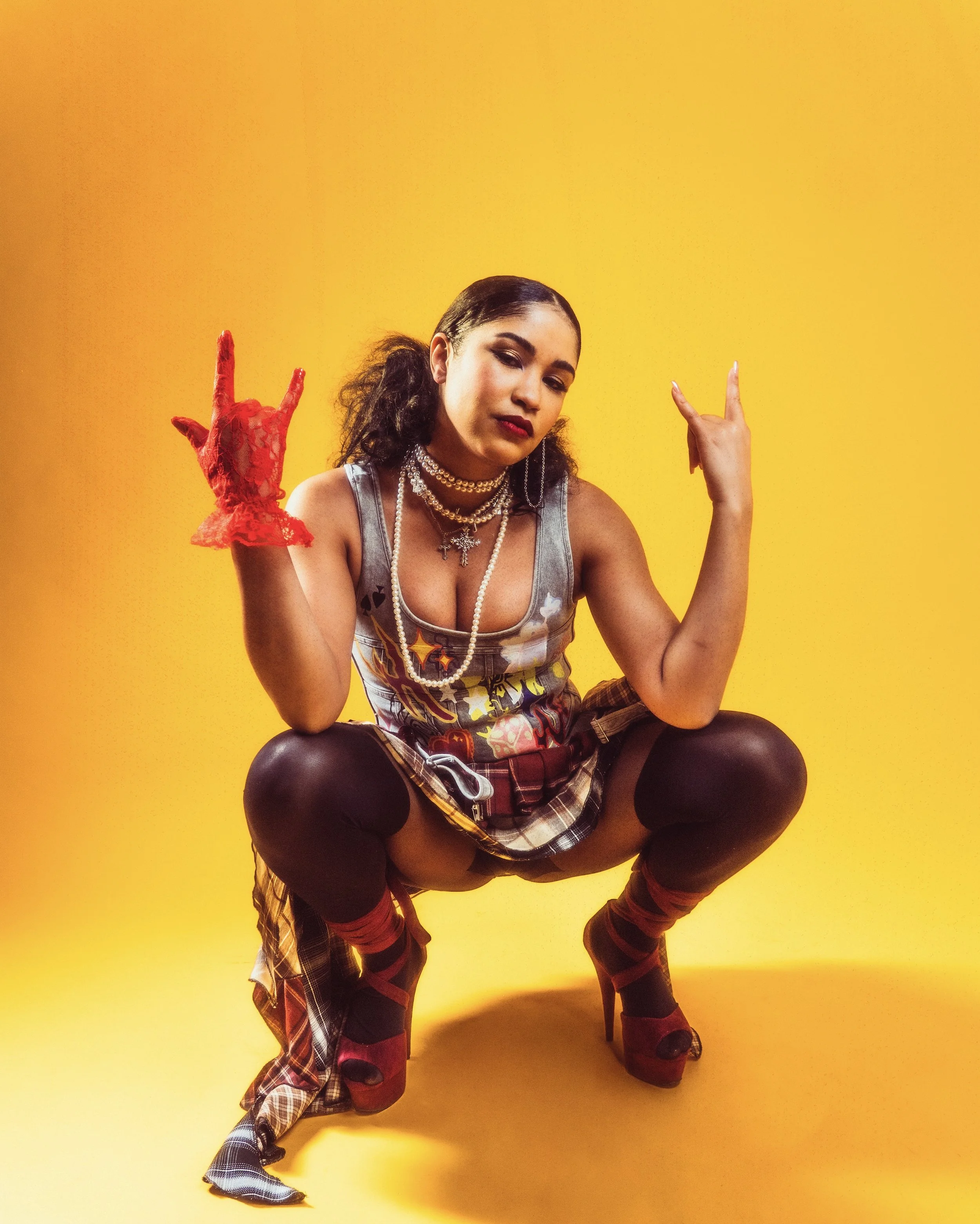 A young woman sitting against a yellow background. She is dressed in a casual, edgy style with layered necklaces, earrings, and makeup. She is making rock hand signs with both hands, wearing a red lace glove on her left hand and red socks with black 