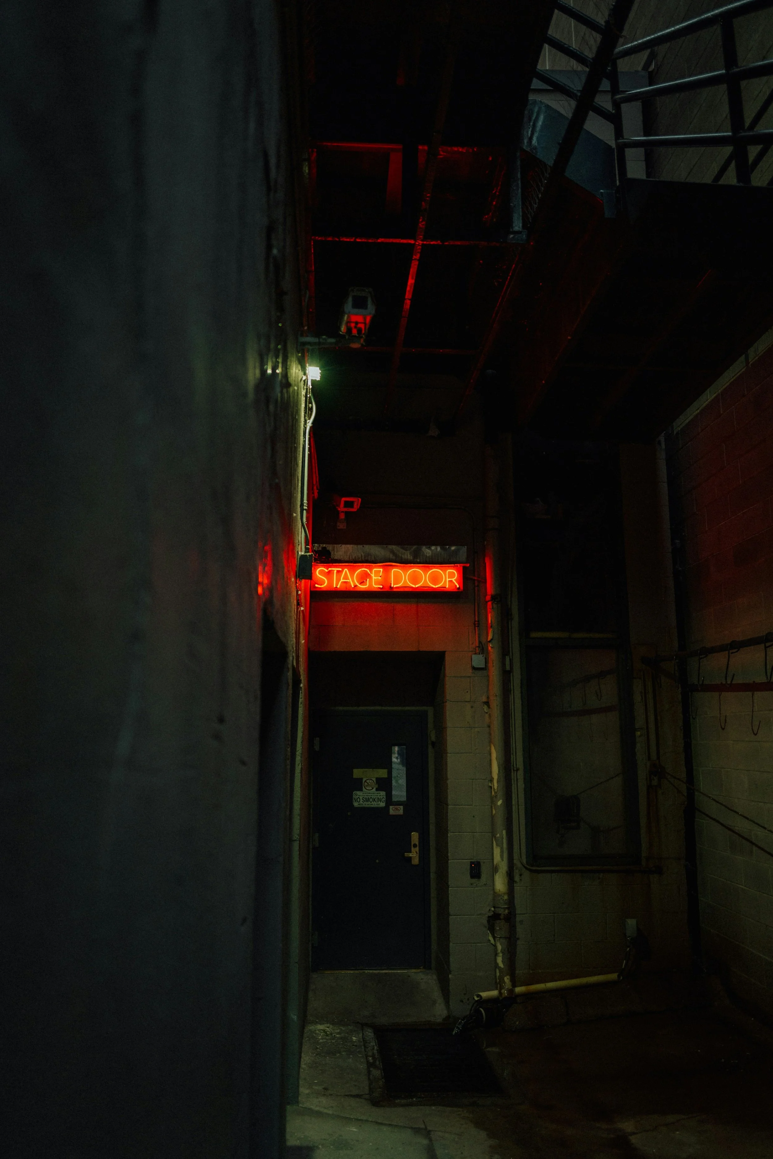 Dark alleyway with a neon red sign reading 'STAGE DOOR' above a black door. The alley has a black metal staircase on the right side and a scooter parked near the door. The area appears to be back entrance of a venue or theater.