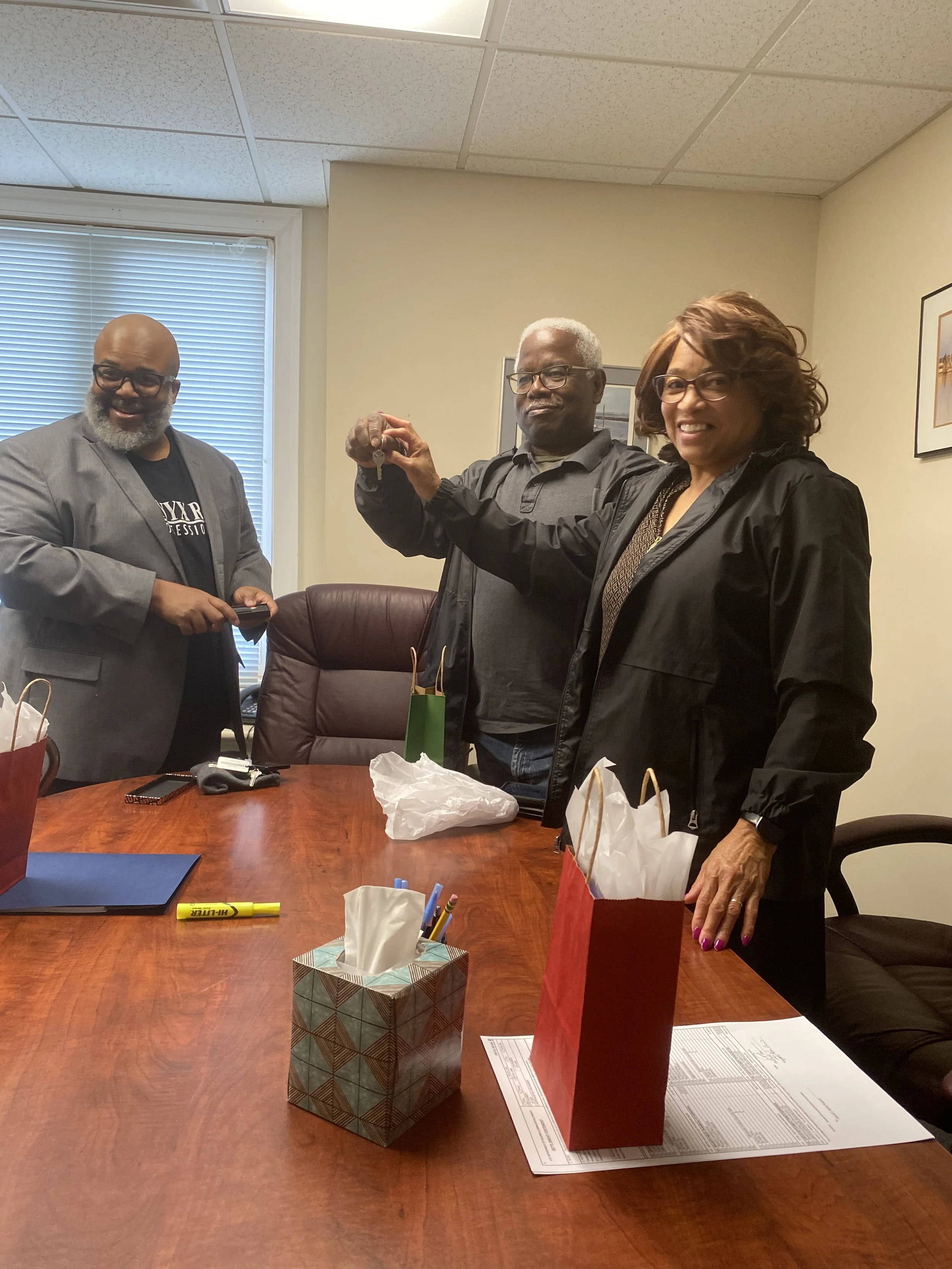 Three adults standing in a conference room. The man on the left is holding a smartphone, the man in the middle is holding a set of keys, and the woman on the right is smiling and extending her hand with a small object. The table has gift bags, tissue boxes, pens, and a yellow highlighter.