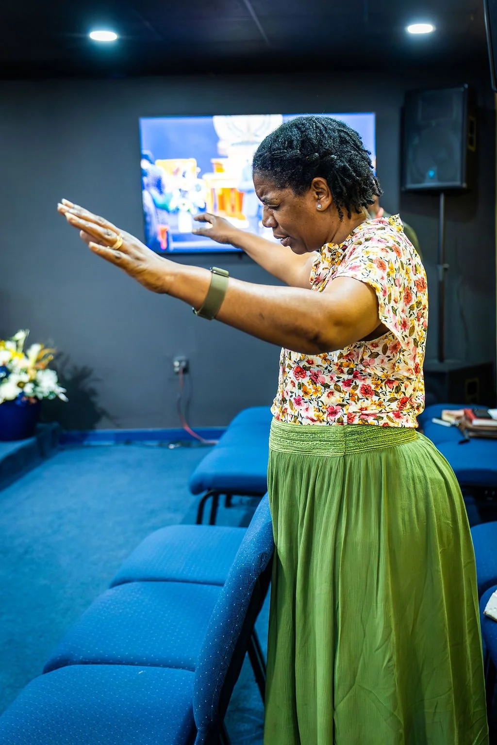 A woman with short dreadlocks and wearing a floral top and green skirt is praying with her arms raised in a church or worship setting.