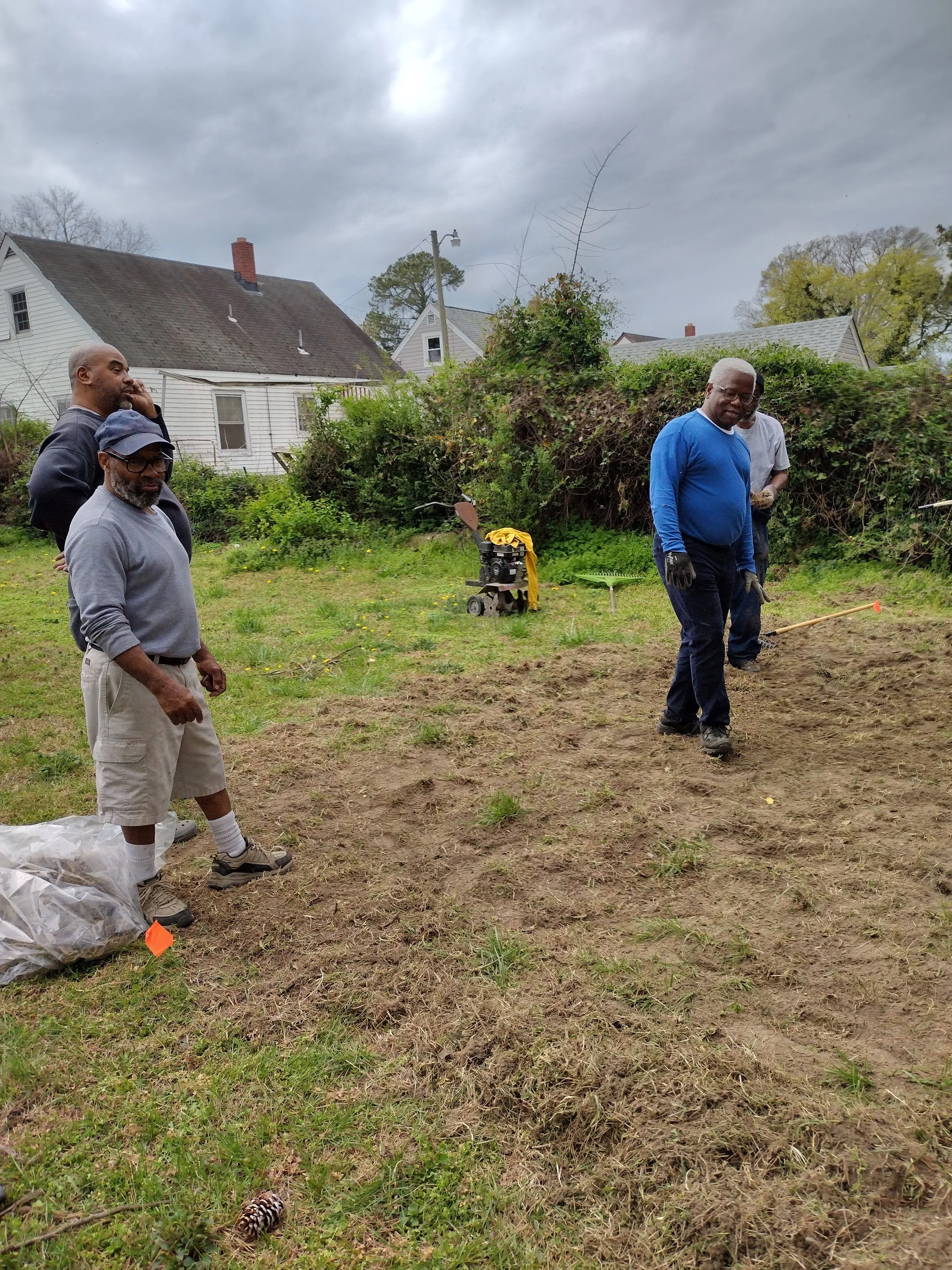 Group of men working on a yard, preparing the soil for planting, with houses and trees in the background.