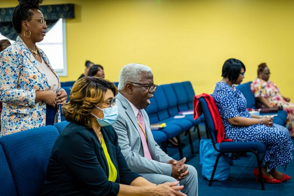 Group of people seated and standing in a room, engaged in a meeting or conference. The woman in the foreground is wearing a face mask, and the man next to her is smiling. The room has a yellow wall and blue chairs.