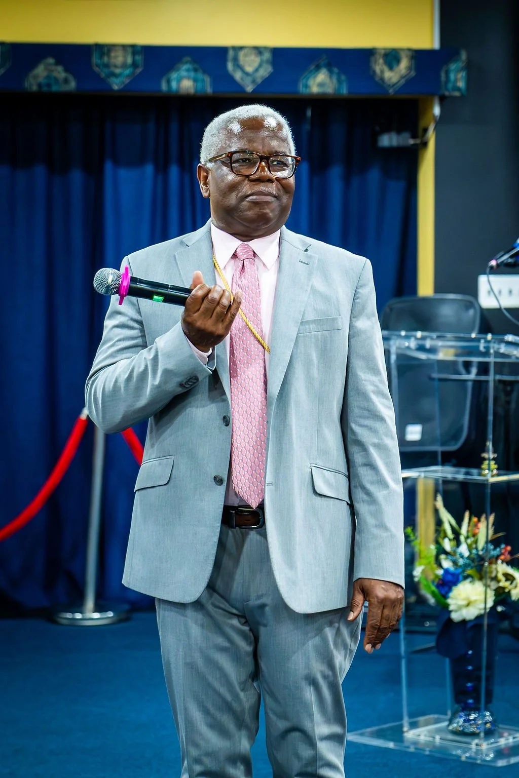 The Bishop, with glasses, dressed in a light gray suit, pink tie, and white shirt, holding a microphone with a pink windscreen, standing in front of a blue curtain and floral arrangements in a formal indoor setting.